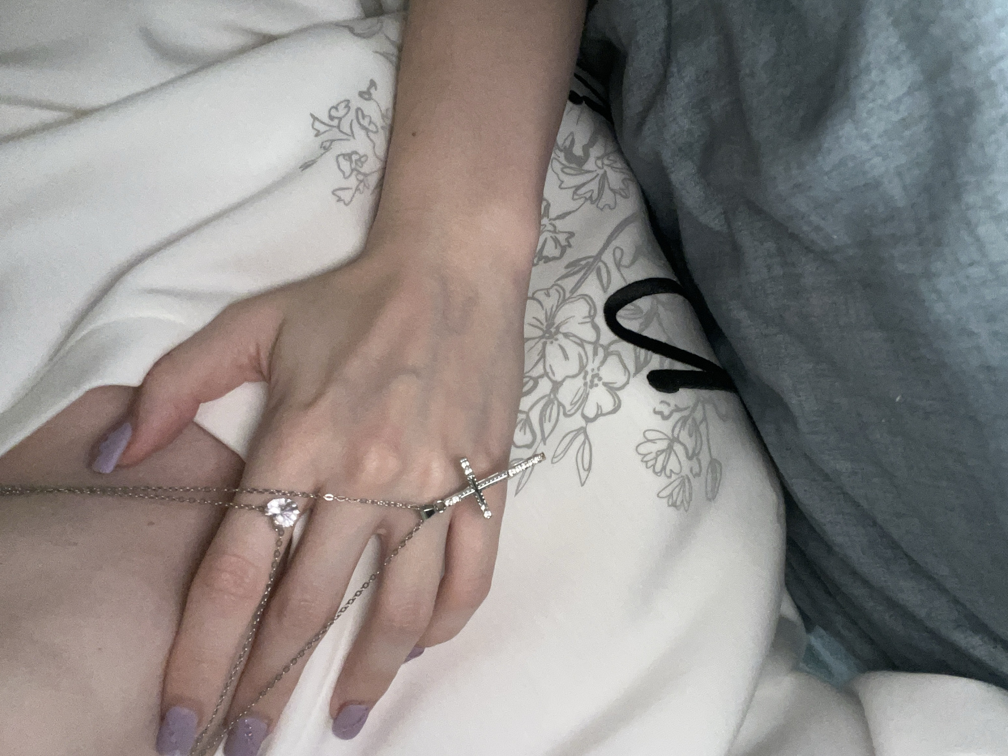 A human hand, painted lavender, adorned with a silver chain accessory that features a cross and a clear gem sits atop floral patterned white bed linens. A gray blanket is positioned on the right of the hand and floral linens. The location is unknown and cannot be determined from the provided image.