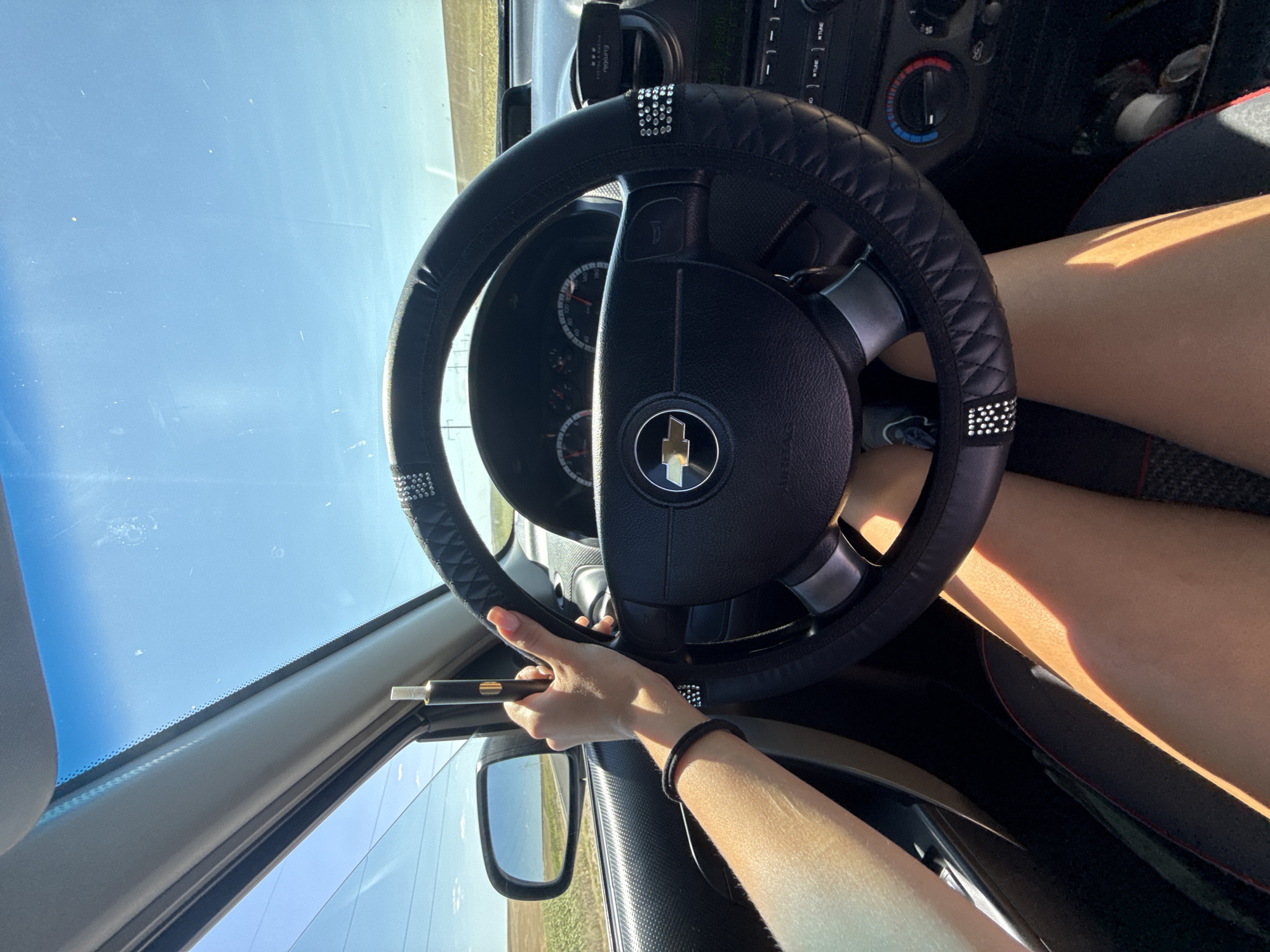 In this close-up, first-person perspective shot from inside a car, a person's hand is shown holding a black vape pen. The hand is positioned near the steering wheel, which is adorned with rhinestones. Sunlight streams through the windshield, illuminating the dashboard and the person's legs. The vehicle appears to be on a rural road, indicated by the dry, grassy landscape visible through the side window. The sky is a clear, bright blue, suggesting a sunny day. The car's interior details include the steering wheel cover, dashboard, air vents, and part of the car's center console. There are no specific landmarks or indicators that place the scene in Cioc-Maidan, Moldova.