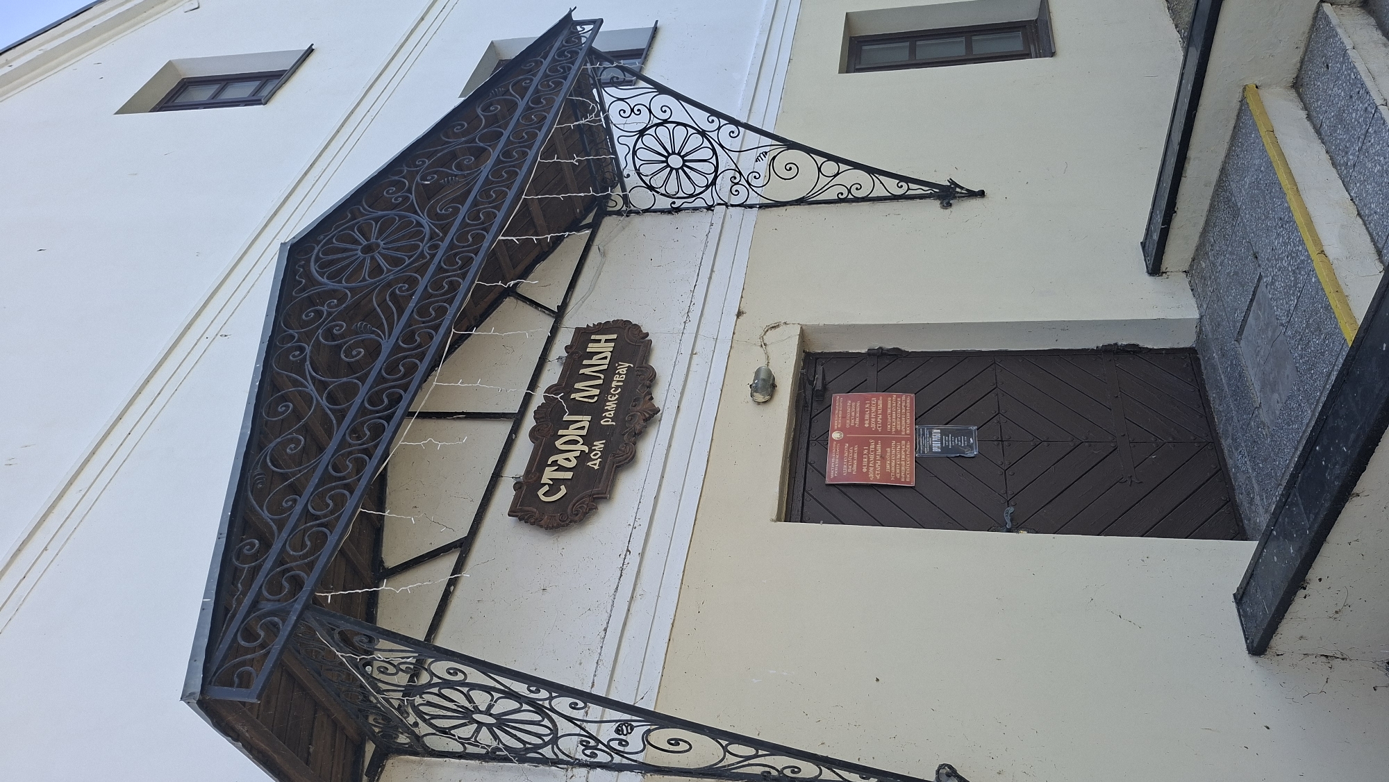 The image shows the entrance to a building labeled in Cyrillic as "СТАРЫ МЛЫН ДОМ РАМЕСТВАУ" which translates to "Old Mill House of Crafts" and is located in Pastavy, Belarus. The building has a cream-colored facade with a dark brown wooden door. Above the door is a decorative dark metal awning with intricate scrollwork and small string lights. A wooden sign with the same Cyrillic text is mounted on the wall above the awning. To the left and right of the entrance are decorative metal brackets supporting the awning, also featuring scrollwork and a circular floral design. There are two small, dark-paned windows visible on the upper part of the building's facade. A set of stone steps leads up to the entrance, with a yellow stripe on the edge of the top step. On the door, there are two notices posted, one of which appears to be an opening hours sign. The overall impression is of a cultural or historical building with traditional architectural elements.