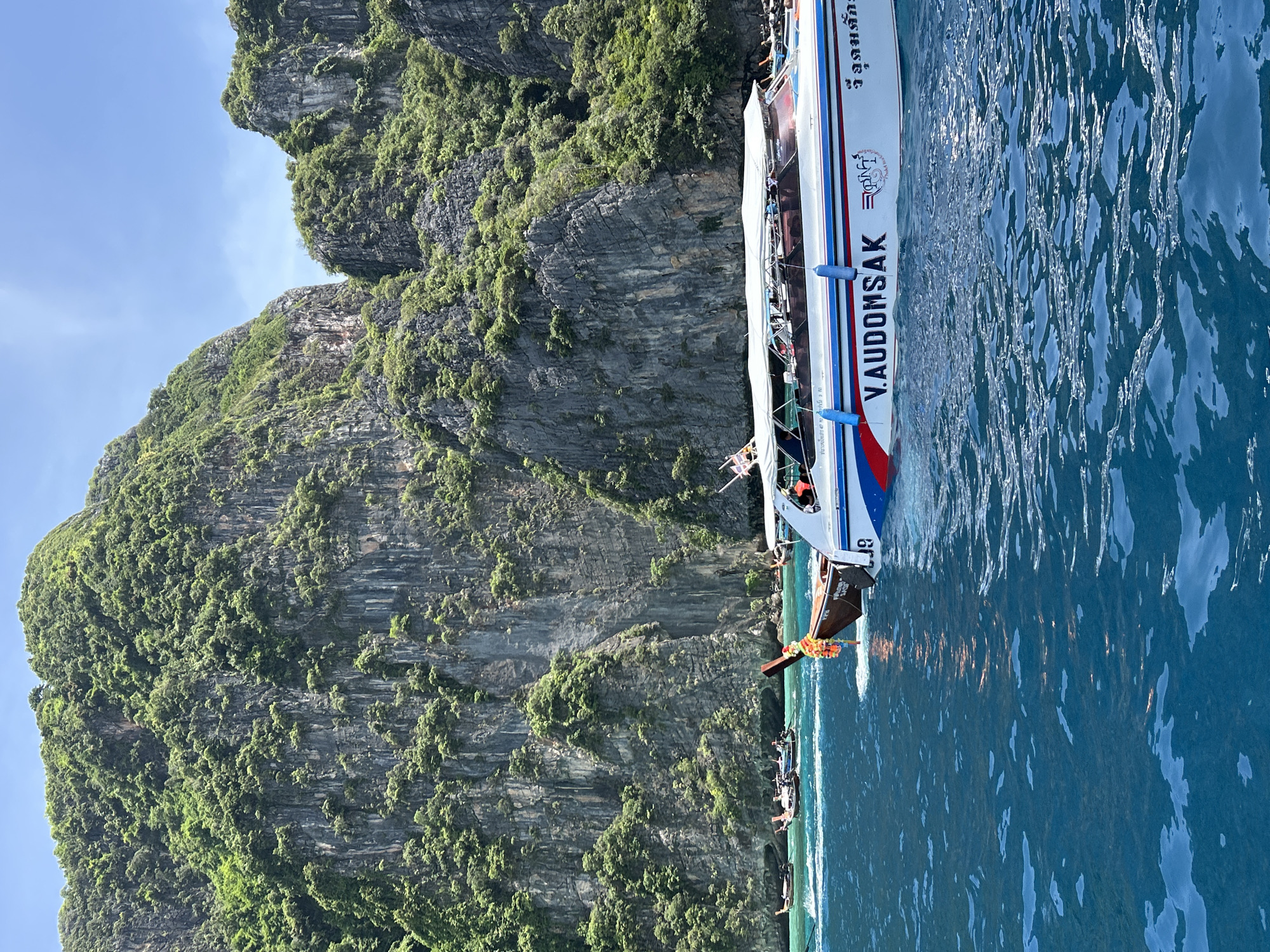 A motorboat with a white canopy and a red, white, and blue stripe sails through turquoise water in front of a lush green cliff face in Ao Nang, Thailand. The boat has the name "V.AUDOMSAK" written on its side. Several longtail boats are visible in the background, further out in the water, near the base of the cliffs. The sky is clear and blue with a few wispy clouds. The water is choppy, creating ripples and reflecting the sunlight. The cliffs are steep and covered in dense vegetation, with a few rocky outcrops visible. The scene depicts a typical tourist activity in a scenic coastal area.