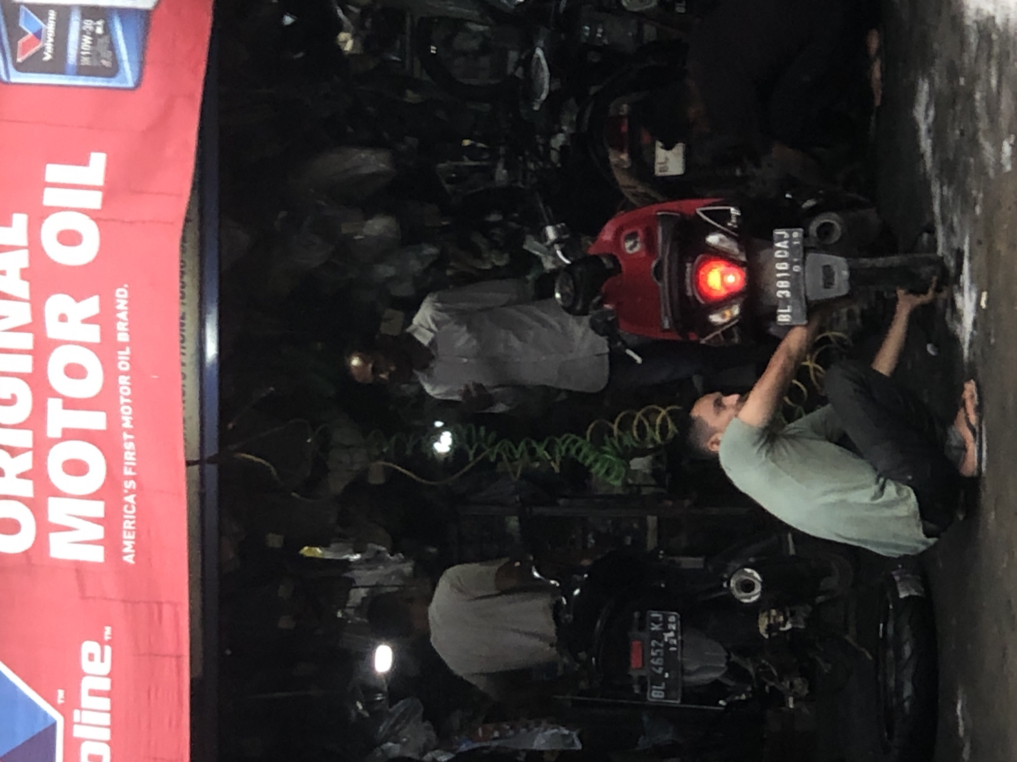 The image depicts a scene inside a dimly lit workshop or store in Titibaro, Indonesia, specializing in motor oil and possibly motorcycle repair. A large red banner with white text announcing "ORIGINAL MOTOR OIL" and "AMERICA'S FIRST MOTOR OIL BRAND" is prominently displayed at the top. Below the banner, several motorcycles are parked, some partially obscured by darkness.
In the foreground, a man wearing a green t-shirt and black pants, along with flip-flops, is crouched down and appears to be working on the rear wheel of a red scooter. The scooter has a license plate that reads "BL 3816 DAJ". To the left, another motorcycle's license plate is visible, reading "BL 4652 KJ".
Further back, a man in a light-colored collared shirt stands near a motorcycle. Behind him, numerous items, likely motorcycle parts or accessories, are stored in an organized or semi-organized manner, creating a cluttered but functional workshop atmosphere. A bright, coiled green hose is also visible, suggesting ongoing work. The floor appears to be concrete and wet in some areas, with white residue present, possibly from cleaning products. The overall impression is of an active, working environment in a commercial establishment.