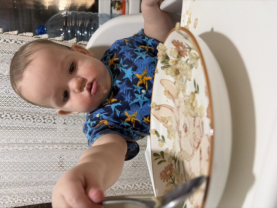 A baby sits in a high chair, wearing a blue star-patterned shirt. His face is turned toward the camera, with food around his mouth. He is holding a spoon in his hand, reaching towards a plate covered in food remnants. The plate has a bunny and floral design. A water bottle sits in the background.