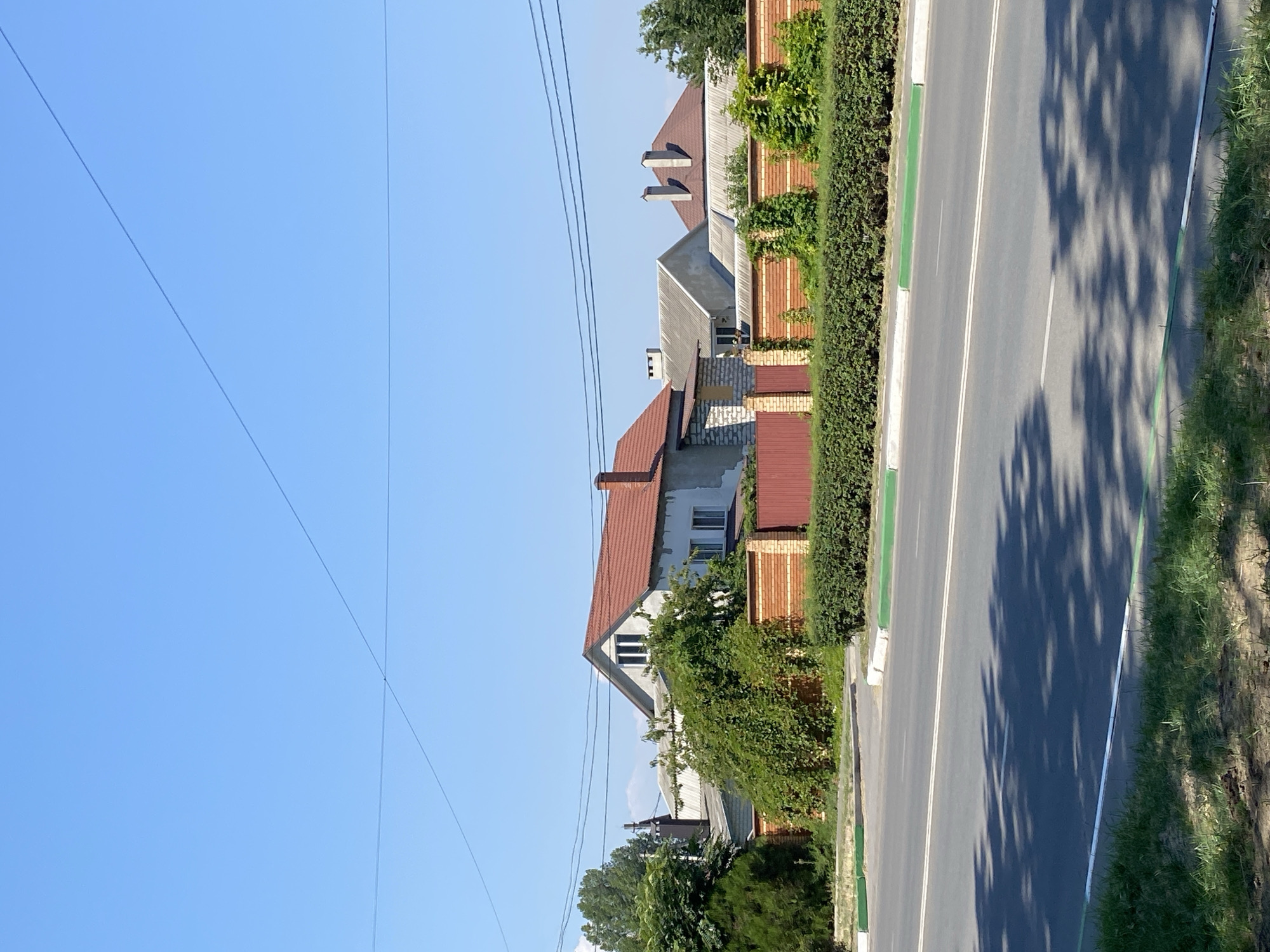 In Chornomorsk, Ukraine, a residential area is depicted on a clear, sunny day. The image focuses on a street in front of several houses. A paved road with white lane markings stretches across the lower half of the frame, bordered by a green and white curb. Shadows of trees are cast across the road. Behind the road, a dense, green hedge runs along a property line. Beyond the hedge, a brick and stone fence encloses several houses. The houses have red tile roofs and are of varying architectural styles. One house has light-colored walls with a prominent red-tiled gable roof and several windows. Another structure behind it has a lighter gray roof. Power lines are visible crisscrossing the bright blue sky. Trees and lush green foliage surround the properties.