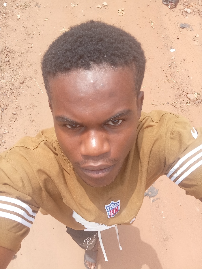 The image displays a dark-skinned male individual from an overhead perspective, situated on reddish-brown, unpaved ground in Nkwelle Ezunaka, Nigeria. The individual has short, dark, tightly curled hair and a discernible mustache and light beard growth. He is wearing a mustard-brown short-sleeved shirt featuring three horizontal white stripes on the upper left arm and a multi-colored NFL shield logo positioned on the center chest. Dark shorts with a white drawstring are visible, along with a single grey slide sandal on his right foot. The subject maintains direct eye contact with the camera, exhibiting a neutral to slightly focused facial expression. The ground surface is irregular, composed of dirt, small stones, and scattered debris.