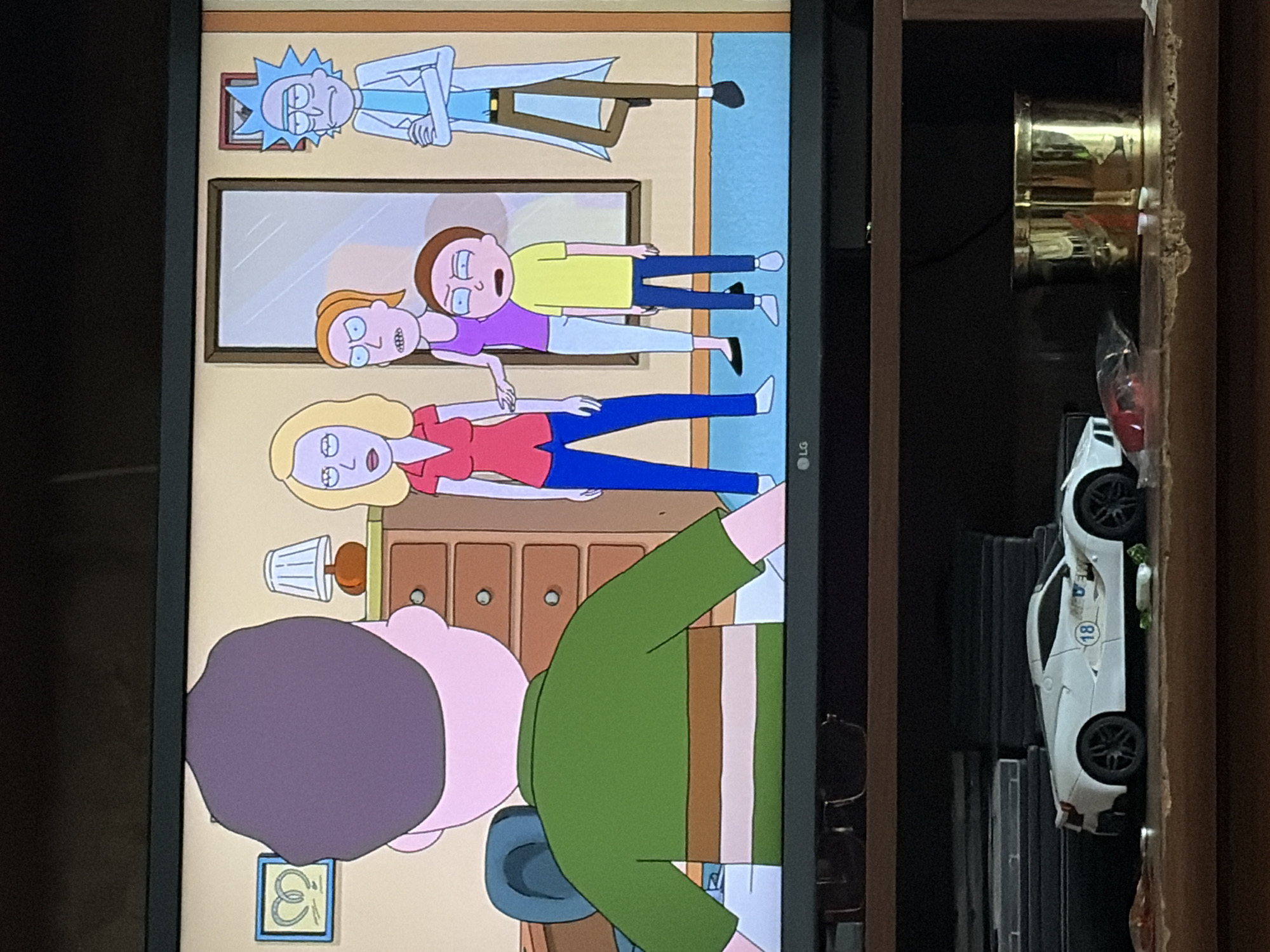 The image shows a television screen displaying an animated scene from the show Rick and Morty. In the animated scene, several characters are gathered in a room. There is a man with a purple head and a green shirt facing away from the camera. To his right, a woman with blonde hair and a red shirt stands next to a man with a blue lab coat and green hair, identified as Rick. Another woman with red hair and a purple tank top stands near a young boy with brown hair and a yellow shirt, identified as Morty. The room appears to be a living room with furniture and a mirror.
Below the television, a shelf holds various items. A white toy car with the number "18" on it is prominently displayed. Next to the car, there are some discarded items, including a red plastic object and a piece of green vegetable. Further to the right, a gold cylindrical container is visible. The overall setting is a domestic environment, with the television displaying a fictional scene. The location is not specified in the image.