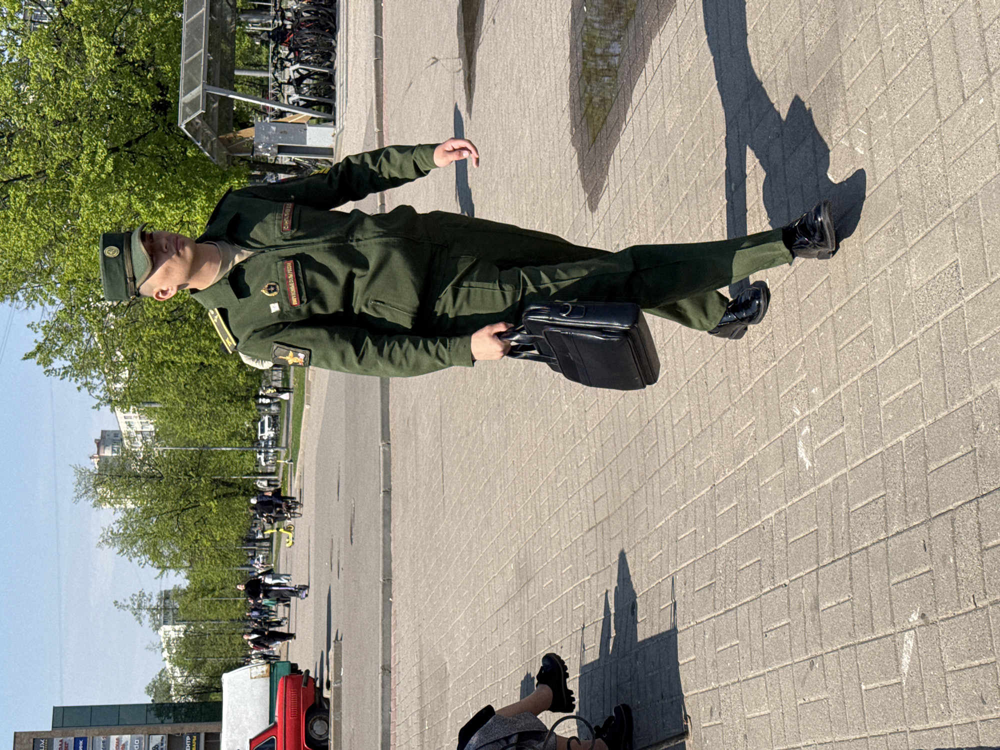 A person in an olive-green uniform and cap, likely a military uniform, is walking on a paved sidewalk. The individual is holding a black briefcase and is wearing black shoes. The location is in Saint Petersburg, Russia, as suggested by text on the uniform. There are trees and buildings visible in the background, along with people on scooters. A bicycle rack can be seen in the upper left, and a red vehicle is visible at the bottom left. The scene appears to be taken on a sunny day, as shadows are visible.