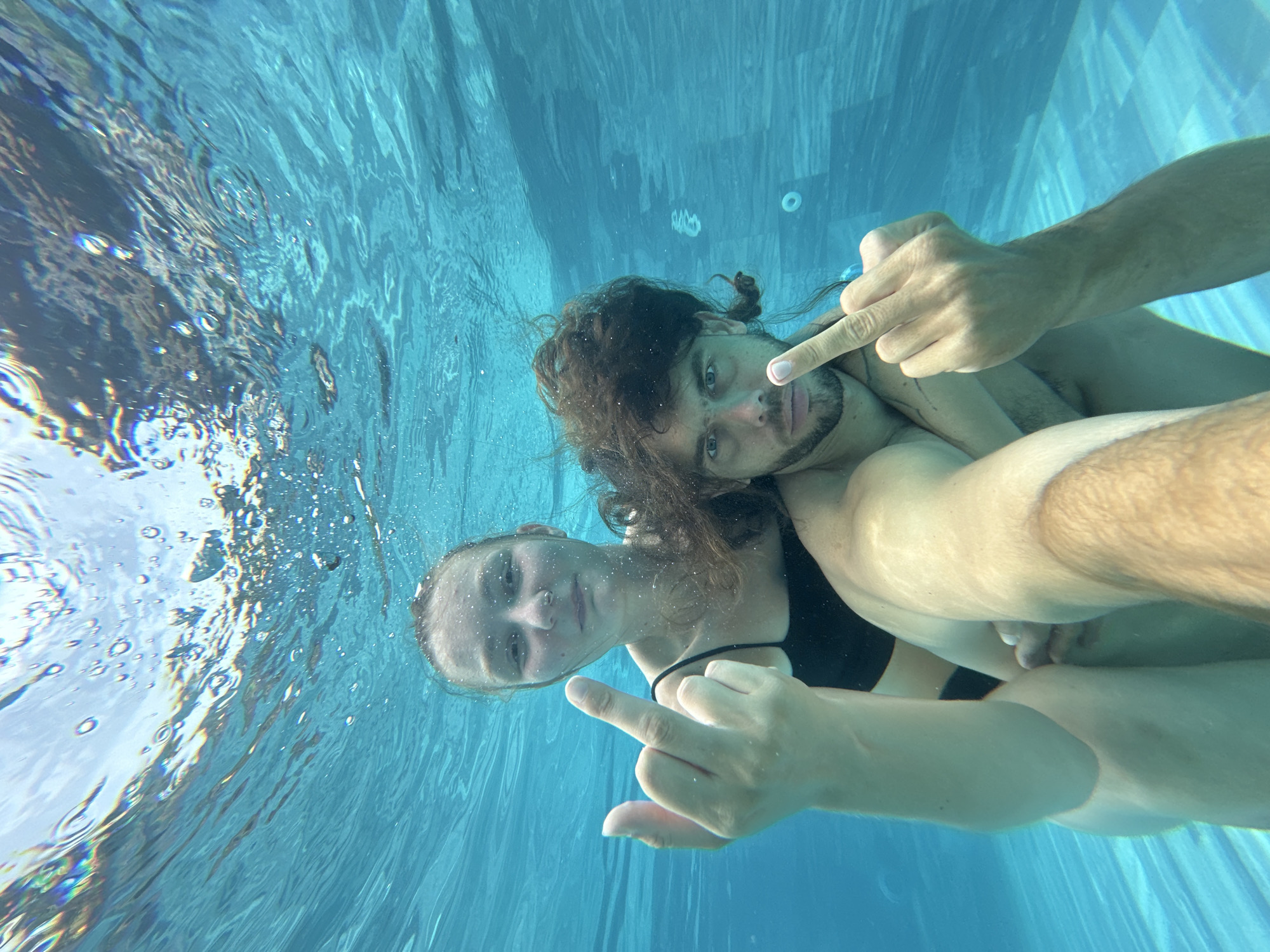 The image depicts an underwater scene in a swimming pool. Two individuals, a woman and a man, are partially submerged. The woman is positioned in front and slightly to the left, wearing a black swimsuit. She is raising her left hand, extending her middle finger. The man is behind her, embracing her, and is also extending his right middle finger. Both individuals have water droplets on their skin and their hair is wet and slightly disheveled due to being underwater. The water is a clear turquoise color, and the pool tiles are visible on the bottom. Bubbles are dispersed throughout the water, and light refracts from the surface, creating a shimmering effect. The overall impression is that of a candid, playful moment captured underwater. The location is specified as Nam Hoa Lư Ward, Vietnam.