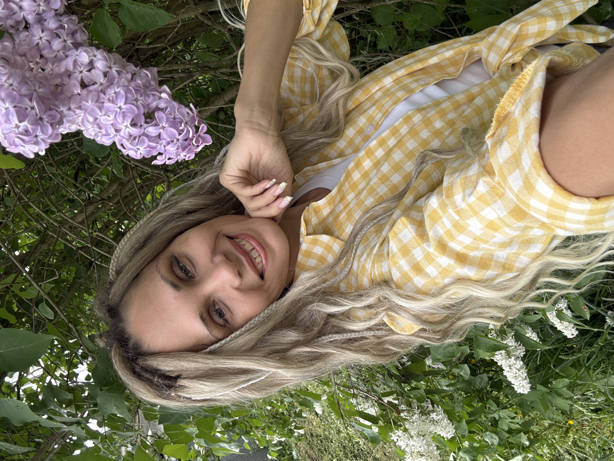 A woman is lying in grass and bushes. She is smiling and looking at the camera. She has long blonde hair with braids and is wearing a yellow and white checkered shirt. There are purple flowers to the left and white flowers to the right of the woman. The location appears to be outdoors in a natural setting, possibly in the city of Rusanovo, Russia.