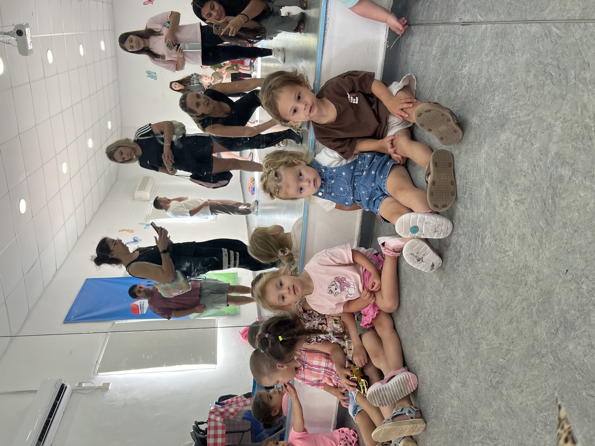 In Kiryat Bialik, Israel, a dance studio with a mirrored wall and a projector is the setting for a group of young children and adults. Five young children are seated on the floor, facing the camera. The child in the foreground on the right wears a brown t-shirt and beige shorts, while the child to their left wears a blue denim romper over a white t-shirt. Another child to the left, wearing a pink t-shirt and pink skirt, is looking to the left. Two more children are seated between the first three, one in a pink and white checkered shirt holding a golden toy car, and another in a patterned dress.
Behind the children, several adults are visible, some interacting with their phones. A woman in a black tank top and black shorts is looking at her phone. Another woman in a pink t-shirt is also holding a phone. In the background, two men and another woman are also present. The studio features a white ceiling with lighting and a projector, a white wall with an air conditioning unit, and a large blue banner with a red and white house graphic. The floor is a gray, textured surface.