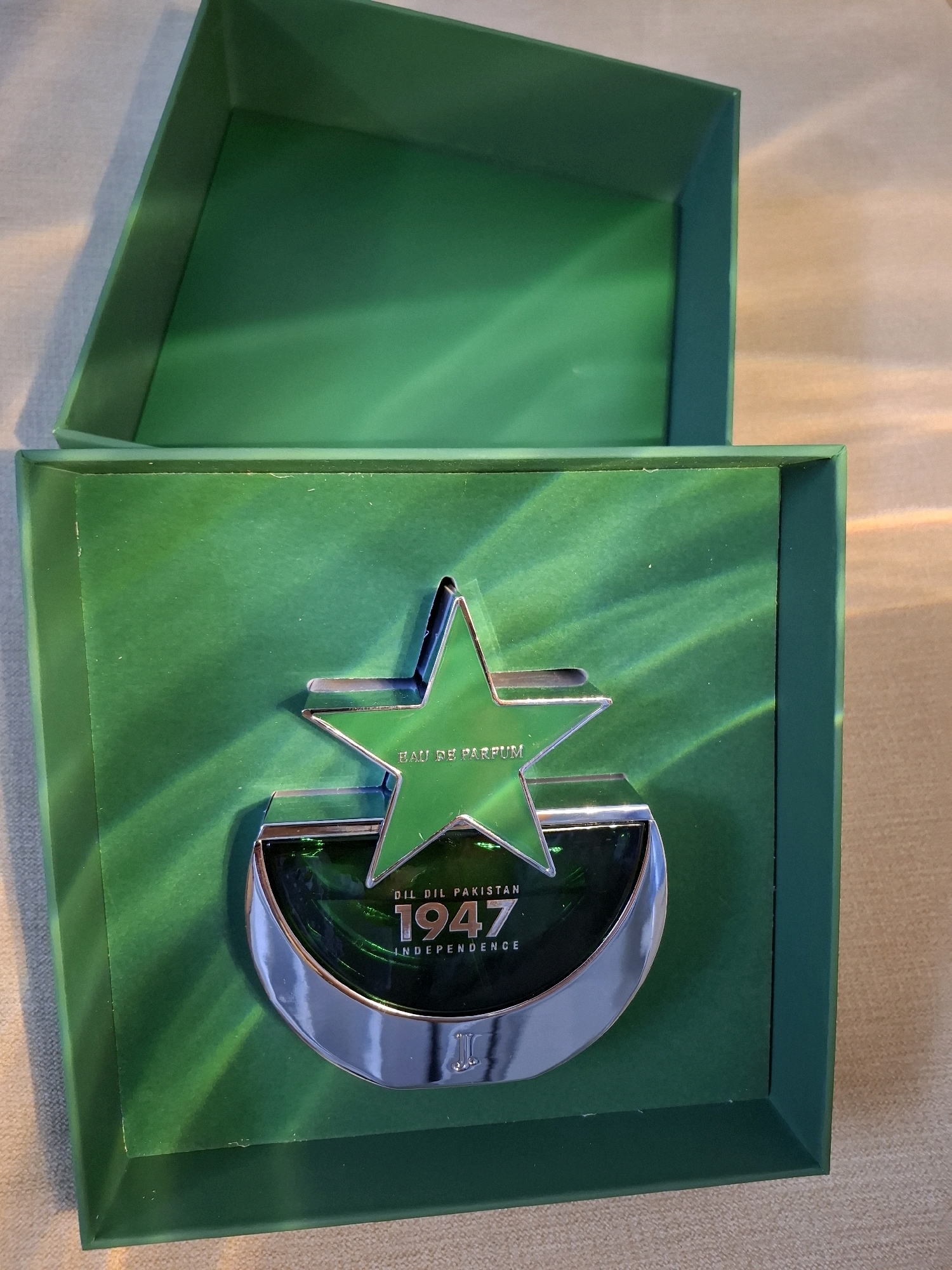 The image shows a green box, open on the left side, containing a perfume bottle. The perfume bottle is shaped like a crescent moon with a star inside. The star has "EAU DE PARFUM" written on it. The crescent moon has "DIL DIL PAKISTAN 1947 INDEPENDENCE" written on it, along with a logo that appears to be an initial. The background of the box is also green with light green stripes. The image is taken from a slightly elevated angle, and the lighting creates a reflective effect on the perfume bottle and the box. There is no discernible location in Abu Dhabi, United Arab Emirates, or any people present in the image.