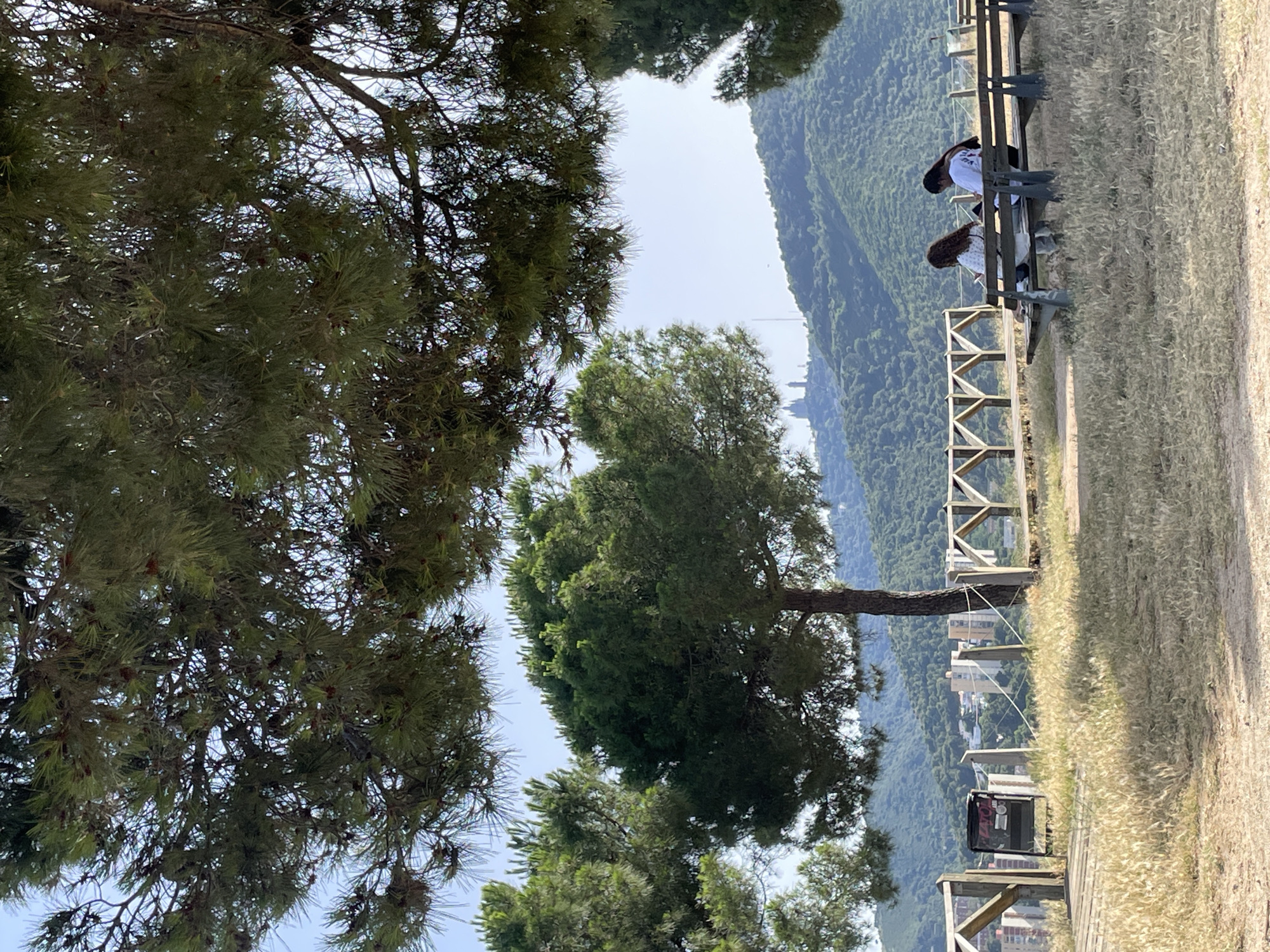 The image depicts a scenic overlook in Barcelona, Spain, offering a panoramic view of the city and surrounding hills. In the foreground, two women sit on a wooden bench, engaged with their devices. The bench is situated on a grassy knoll, with dry, golden grass indicating a warm climate. Behind them, a wooden fence lines the edge of the overlook. Beyond the fence, several buildings, including apartment complexes, are visible against the backdrop of lush green hills. Dominating the distant skyline is a prominent religious building, possibly a church or monastery, situated atop one of the hills. The sky is clear and blue, suggesting a sunny day. The overall scene conveys a sense of tranquility and natural beauty within an urban landscape.