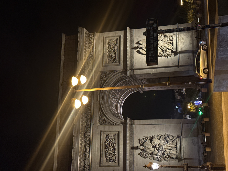 The image depicts a nighttime view of the Arc de Triomphe, a prominent neoclassical triumphal arch, situated in Paris, France, at the center of Place Charles de Gaulle (formerly Place de l'Étoile). The monumental structure, constructed of light-toned stone, is illuminated, revealing its detailed sculptural reliefs, including "Departure of the Volunteers of 1792" on the left and "The Triumph of 1810" on the right.
A tall, four-light street lamp, featuring spherical luminaires emitting a bright yellow glow and pronounced lens flares, is positioned centrally in the foreground. To the far left, a traditional lantern-style street lamp is partially visible. On the right side of the frame, a dark, vertical street sign pole bears the white inscription "ARC DE TRIOMPHE."
A light-colored, possibly silver, station wagon or small SUV is visible on the paved street in the lower right quadrant, with its headlights illuminated. The ground in the immediate foreground is paved, delineated by dark, short bollards or barriers. Through the main archway, distant city lights, including elements of green and blue illumination, along with faint outlines of structures and trees, are discernible against the dark night sky. No individuals are visible in the depicted scene.