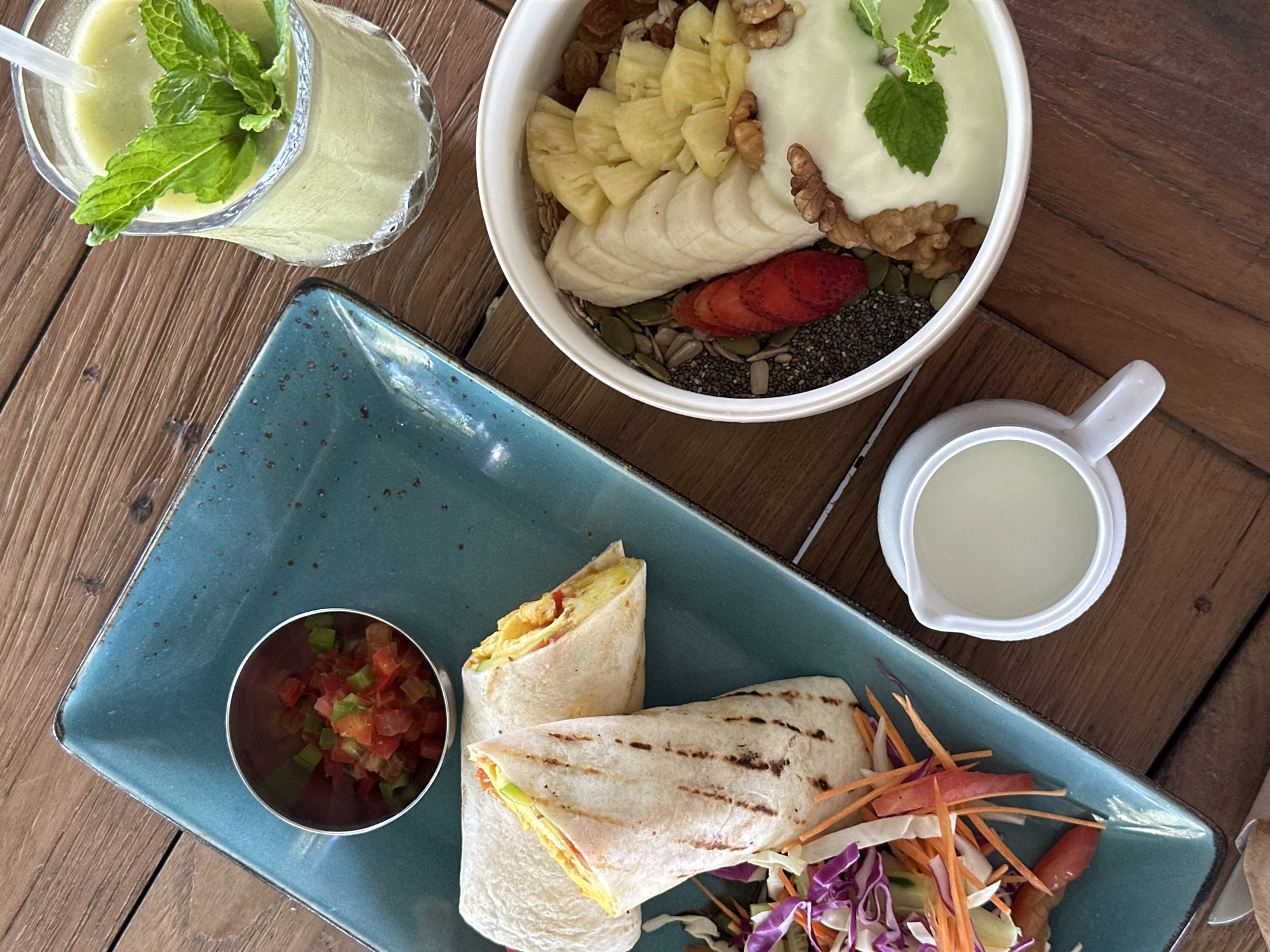 An overhead shot on a wooden table shows a meal including a smoothie, a breakfast bowl, and two breakfast burritos on a teal-colored plate with a side of pico de gallo and salad. A white creamer is also on the table. The meal may be located in the city of Pekutatan, Indonesia.