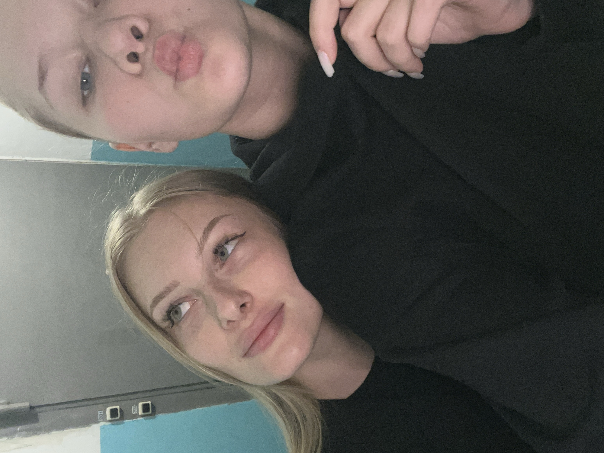 Two young women are captured in a close-up, selfie-style photograph. The woman on the left is positioned slightly behind and to the left of the woman on the right. She has blonde hair, is wearing a black top, and rests her head on the shoulder of the other woman. Her eyes are looking upwards and to the left. The woman on the right, closer to the camera, has her head tilted forward and is making a kissing face. She has blonde hair pulled back and is wearing a black hoodie. Her right hand, with long light-colored fingernails, is visible near her chest, resting on the hoodie.
The background appears to be an indoor setting. To the left, a wall is visible with vertical blue and white paint sections. On the gray surface of this wall, two small, dark rectangular labels with white numbers are affixed. The upper label reads "201" and the lower label reads "200". This labeling suggests the location could be a hallway in a building, possibly an educational institution or residential building, consistent with locations found in the city of Perm, Russia. The lighting is dim and appears to come from the front, casting soft shadows.