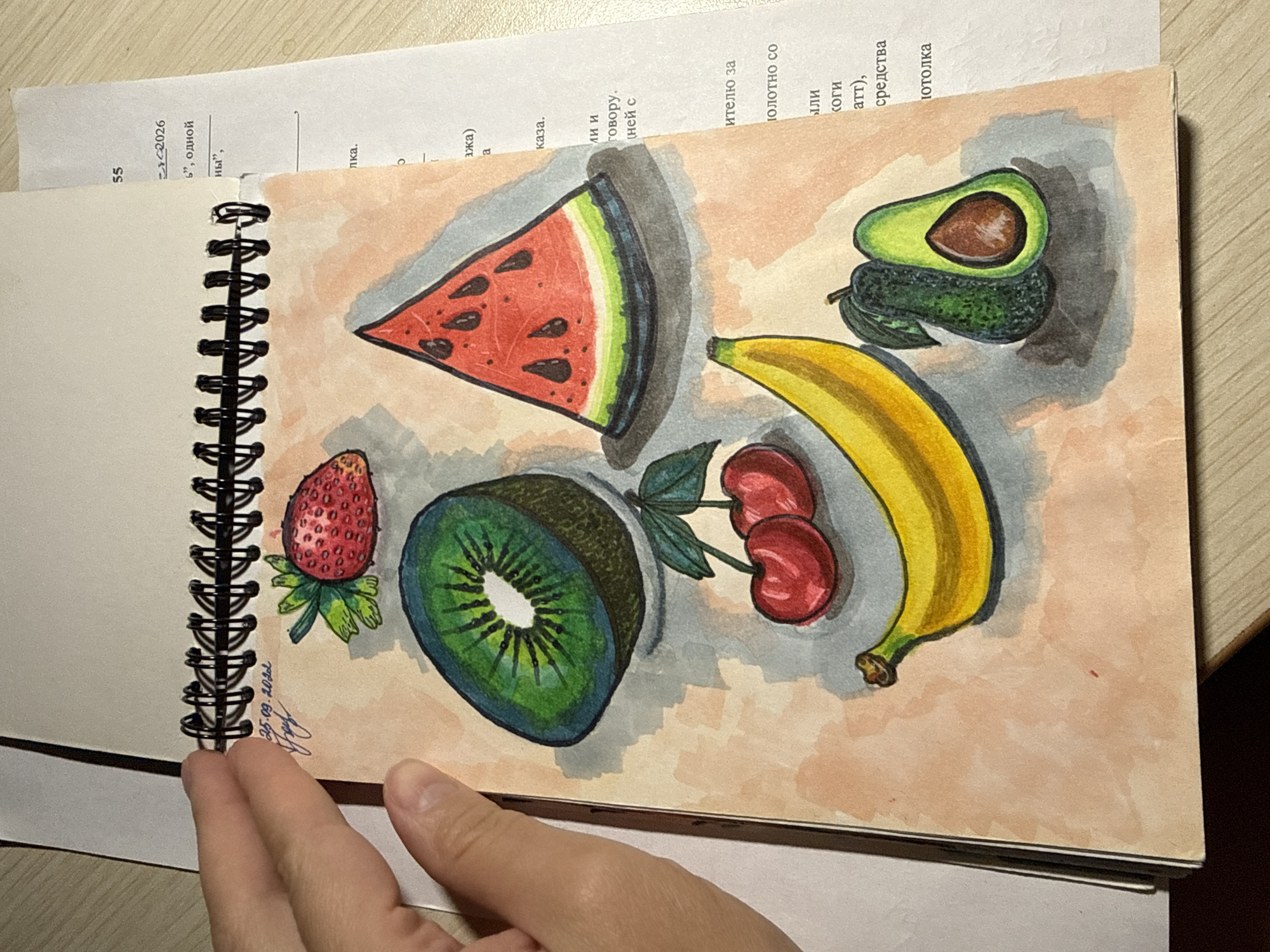 The image shows a close-up of a sketchbook page featuring a colorful drawing of various fruits, including a strawberry, a kiwi slice, a watermelon slice, cherries, a banana, and an avocado. The drawing is done in a vibrant, illustrative style with what appears to be watercolor or colored pencils, and shadows are rendered with gray marker. The sketchbook is open, with a hand visible on the left side, holding the pages. In the background, parts of a document with text in Russian are visible, suggesting the sketchbook is placed on a desk or table. The overall scene is indoors, with the lighting suggesting an office or study environment. There are no discernible elements that would indicate a specific city or location.