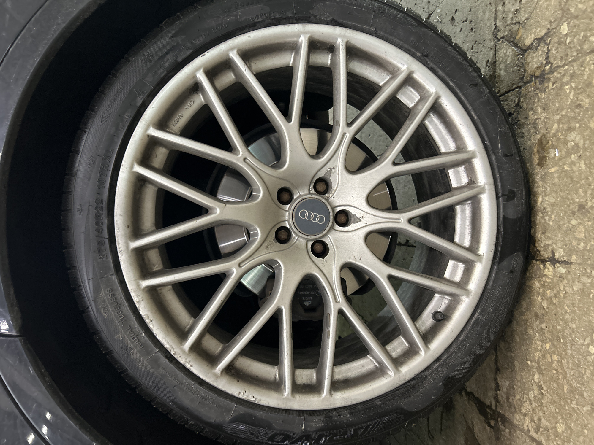 A close-up, eye-level shot shows a silver Audi wheel with a black tire. The wheel is multi-spoke and shows signs of wear and dirt. The tire has text on its sidewall, including "RADIAL TUBELESS" and "275/40R22 107H XL". The Audi logo is visible in the center of the wheel. The tire is mounted on a vehicle that is parked on a wet, concrete surface. The background is dark and out of focus, but appears to be part of a car's wheel well. There are no people visible in the image. The scene is likely in Tallinn, Estonia, given the presence of the Audi brand which is common in European cities, and the overall urban setting suggested by the concrete.