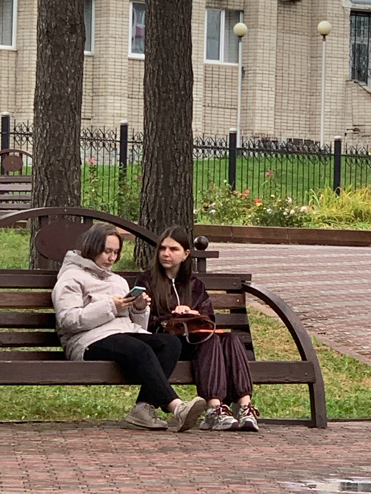 Two young females sit on a park bench. The female on the left, with short brown hair, wears a light-colored puffer jacket and black pants. She is looking down at her phone. The female on the right, with long dark hair, wears a dark tracksuit and is looking to her left. A brown handbag rests on the bench between them. The bench is made of wood and has a curved, dark metal frame. Behind the bench is a grassy area with bushes and flowers, followed by a black metal fence and a brick building with windows. The ground in front of the bench is paved with reddish-brown bricks. The image appears to have been taken outdoors in Prokopyevsk, Russia.