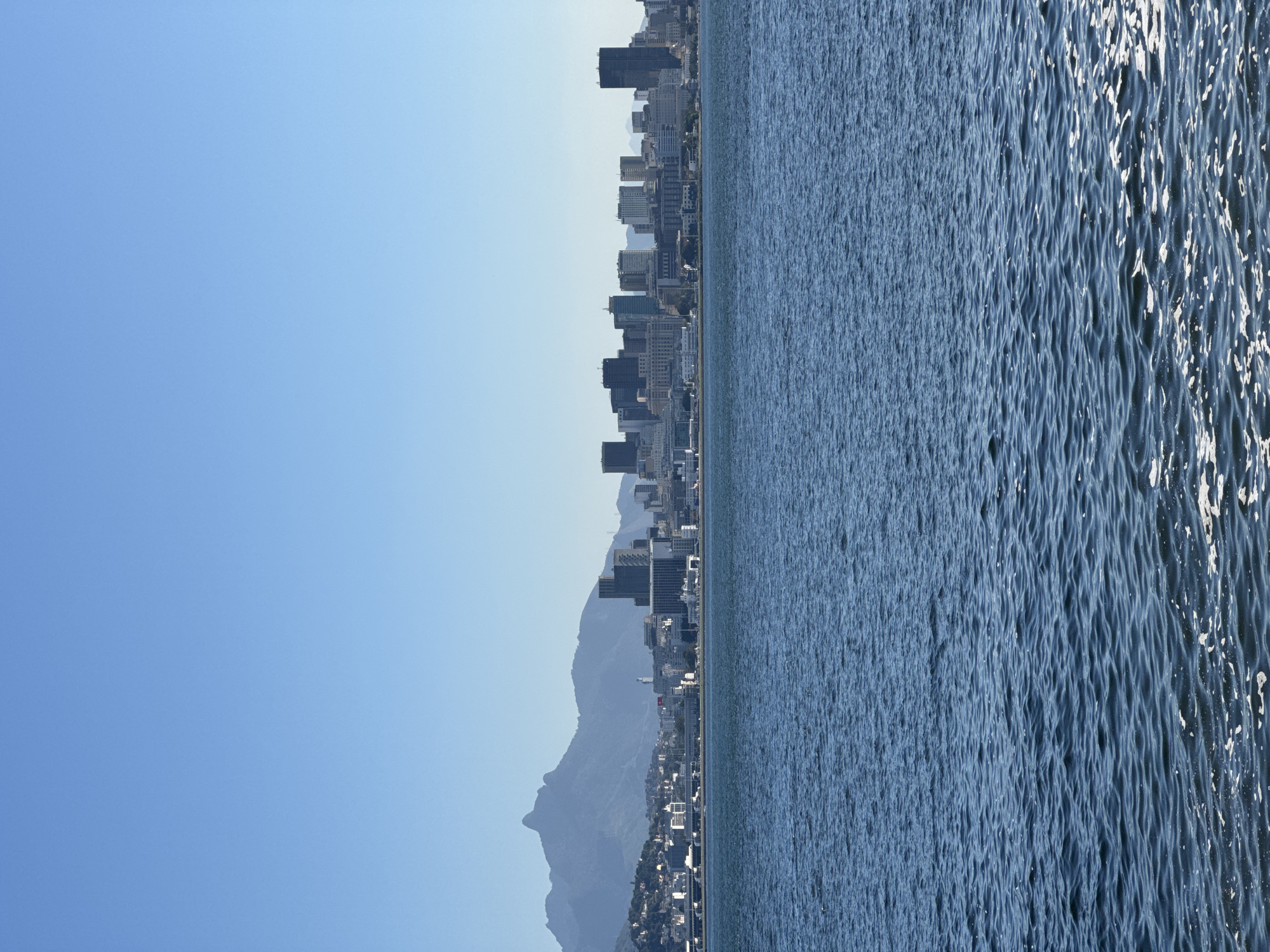 This is a wide shot of the city of Niterói, Brazil, taken from the water. The image features the city skyline in the background, with a range of buildings from low-rise structures to tall skyscrapers. Behind the city, a range of mountains are visible, with one prominent peak having a distinctive shape. The foreground is dominated by a large body of water, likely Guanabara Bay, with gentle waves creating a textured surface. The sky is a clear, bright blue. The overall scene depicts a coastal urban landscape under a sunny sky.