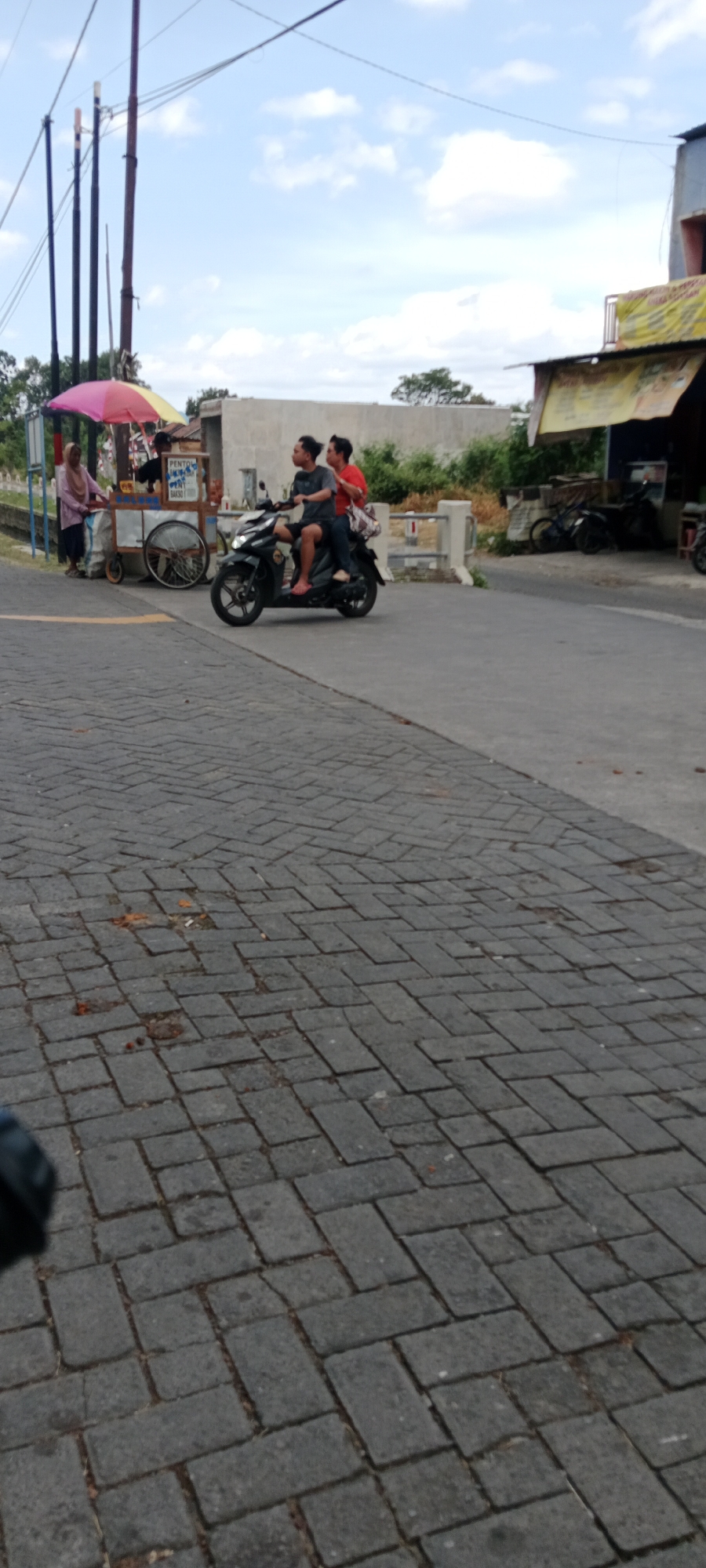 In Maguwoharjo, Indonesia, a street scene unfolds with two individuals riding a black motorcycle. The rider, a young man with dark hair, wears a gray t-shirt and dark shorts. The passenger behind him, also a young man with dark hair, is dressed in a red t-shirt and dark shorts. They are both looking to their right, seemingly observing something off-camera. To the left of the motorcycle, a food cart is visible, adorned with a colorful umbrella. A woman in a light-colored headscarf and tunic stands near the cart, interacting with it. The ground is paved with interlocking rectangular bricks, and a paved road runs alongside it. In the background, a low concrete wall separates the road from a patch of greenery and trees, with a building also visible in the distance. The sky above is partly cloudy, with scattered white clouds against a blue expanse. The overall scene depicts a typical street view in a semi-urban area.