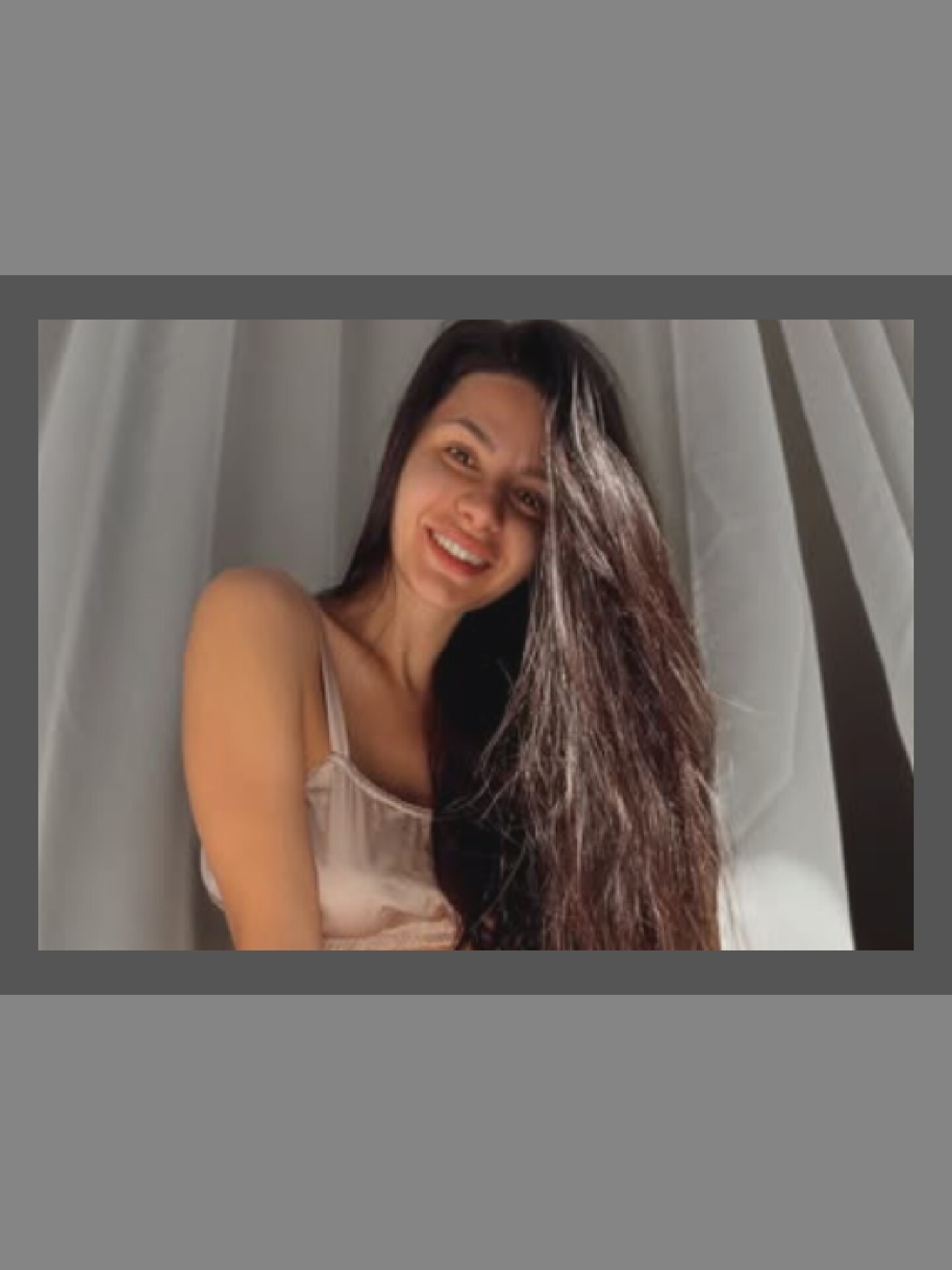 A woman with long, dark hair smiles at the camera. She is wearing a light-colored slip and is positioned in front of a light-colored curtain. The lighting suggests natural light, possibly from a window. The background is out of focus, with the woman being the clear subject.
