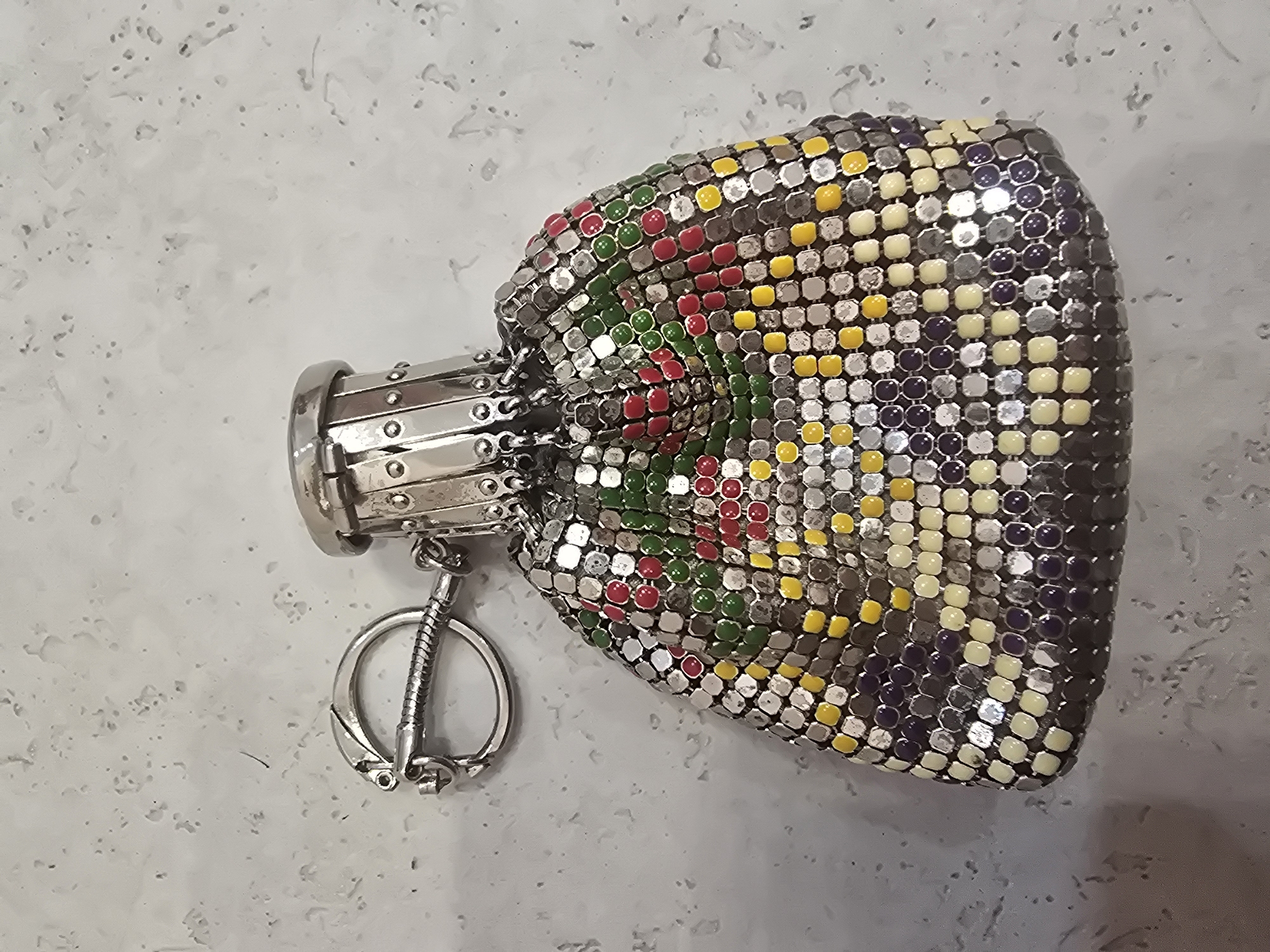 A mesh drawstring pouch, attached to a keychain with a decorative clasp, rests on a light gray, speckled surface. The pouch is constructed from interlocking metal scales, with colorful accents in red, green, yellow, and purple. The drawstring and clasp mechanism at the top of the pouch are silver-toned metal. The background appears to be a textured countertop or floor. There is no discernible urban context or specific location within Ulan-Ude, Russia.