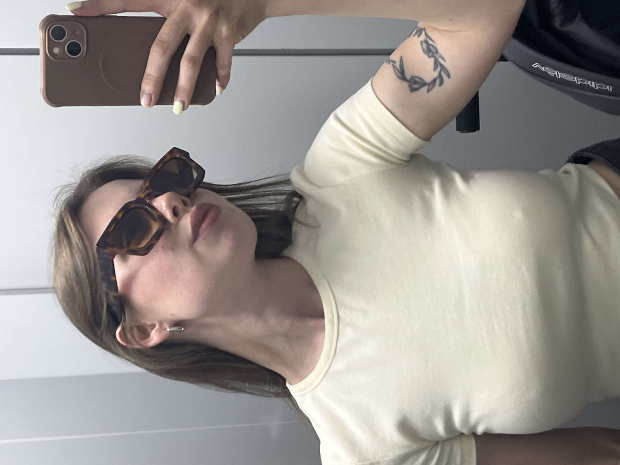 The image captures a woman taking a mirror selfie indoors in Chaikovsky, Russia. She holds a brown smartphone, likely an iPhone, in her right hand, which has light yellow polished fingernails. The woman's head is tilted, and she is wearing large, dark brown tortoiseshell-patterned sunglasses. Her lips are slightly puckered. She has long, dark hair and a small silver hoop earring in her left ear. She is dressed in a plain, light yellow, short-sleeved t-shirt. A tattoo of a circular wreath with leaves and small buds is visible on her right upper arm. In the background, to the right, a dark object, possibly part of a stroller or car seat, is partially visible, bearing the faintly discernible text "joolz." The setting appears to be a simple indoor space with a plain, light-colored wall or reflective surface. The activity is a casual self-portrait.