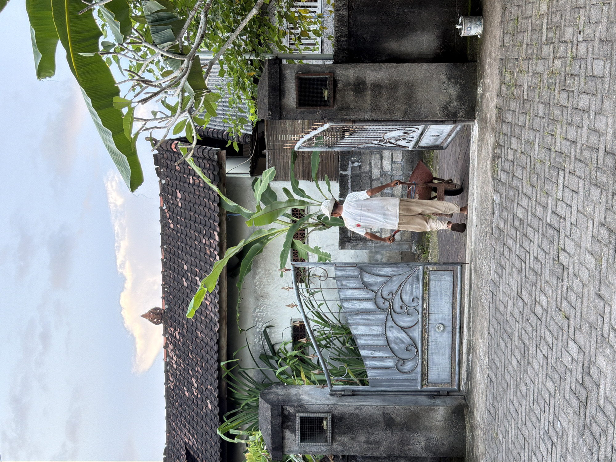 An eye-level, exterior shot shows a man walking along a brick path in Kuta Selatan, Indonesia. He is wearing a white shirt, khaki pants, brown shoes, and a white hat. He is pushing a rusty, cone-shaped handcart. To the left of the man is a building with a tile roof. A banana tree and other tropical plants are near the building. There is an ornate metal gate in front of the building. The walkway is made of gray brick. The sky is visible in the upper left corner.
