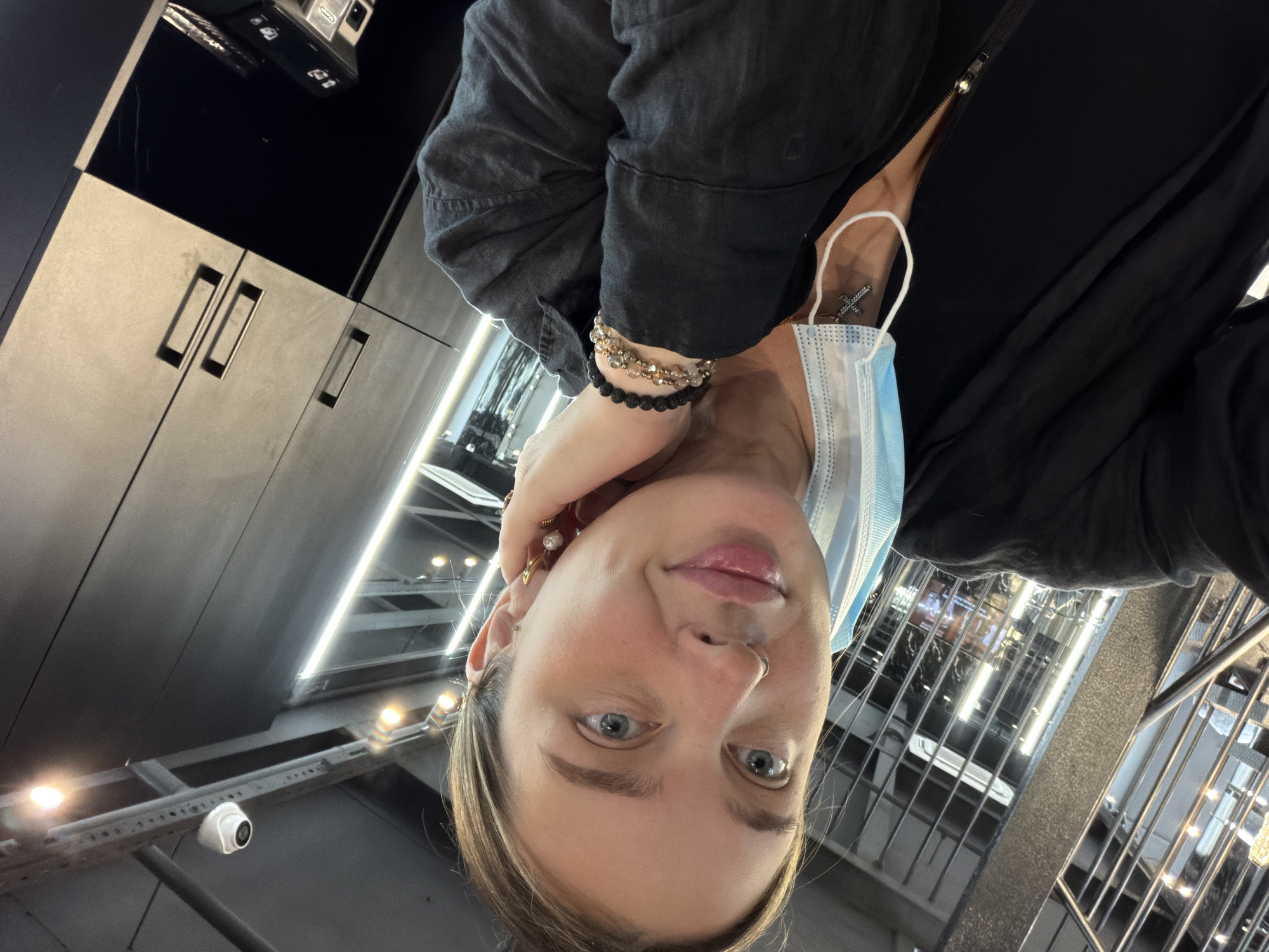 The image shows a woman wearing a black jacket and a light blue surgical mask. She has multiple bracelets on her left wrist and earrings. The background includes what appears to be a modern interior with glossy cabinets, track lighting, and elements suggestive of a public or commercial space. Given the presence of surveillance cameras and the overall architecture, the location may be a retail store, office building, or public transportation hub. It's difficult to confirm the location in Moscow, Russia, without additional visual cues or references.