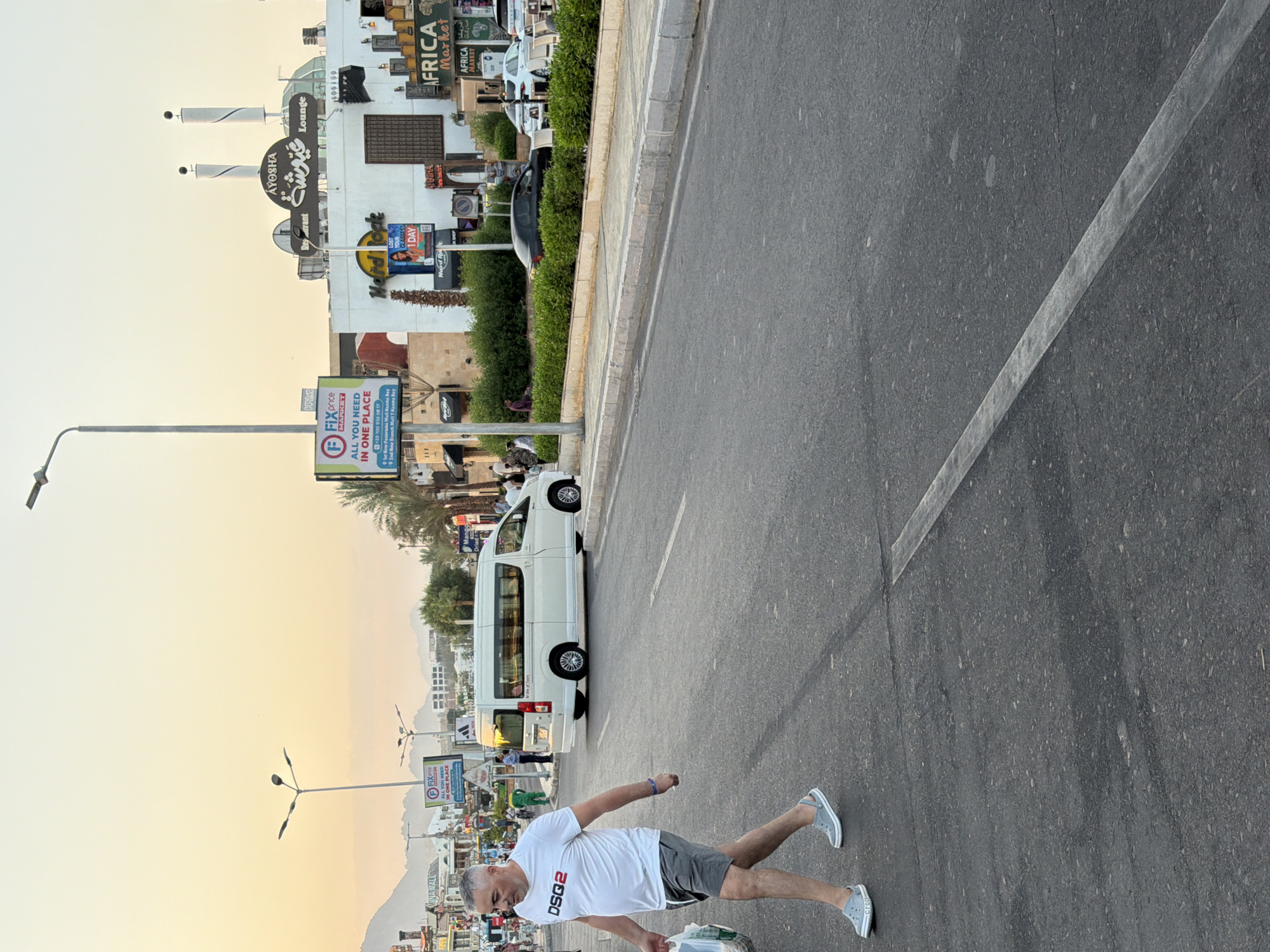 In Sharm El Sheikh, Egypt, an adult Caucasian male with graying hair, wearing a white "DSQ2" t-shirt and gray shorts, walks across a paved road. He is carrying a plastic bag in his left hand and wears gray Croc-style shoes. Behind him, a white minivan is parked on the right side of the road. The scene appears to be a commercial street with buildings, shops, palm trees, and streetlights. The sky is a warm, pale orange, suggesting either sunrise or sunset. In the background, mountains are visible. A prominent sign for "AYOSHA Lounge" with Arabic script is on one of the buildings. Other signs indicate businesses like "Hard Rock Cafe" and "AFRICA Market". The road has a white line marking.
