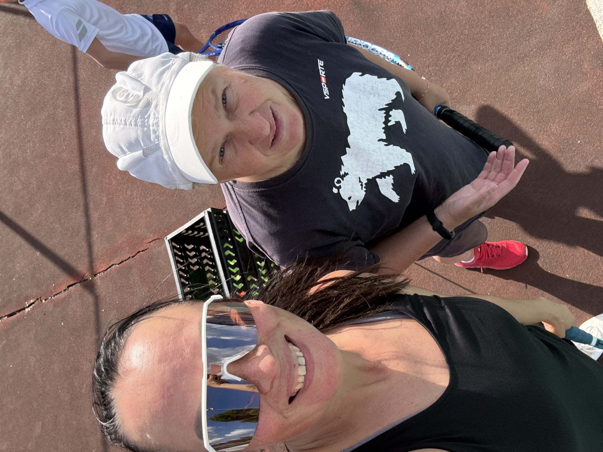 The image shows a top-down view of two individuals on a red, cracked outdoor surface, likely a tennis court, given the presence of tennis rackets and a ball crate. The person in the foreground on the left is a woman with dark hair, wearing large reflective sunglasses, a black tank top, and a tennis racket. She is smiling. To her right, a person wearing a white cap, a dark t-shirt with a white bear graphic, and red tennis shoes is also visible, holding a tennis racket. In the background, another person in a white polo shirt and dark shorts can be seen. The environment appears to be a sunny day, with shadows cast on the court. The presence of a ball crate suggests a tennis match or practice. The location is identified as Chaikovsky, Russia.