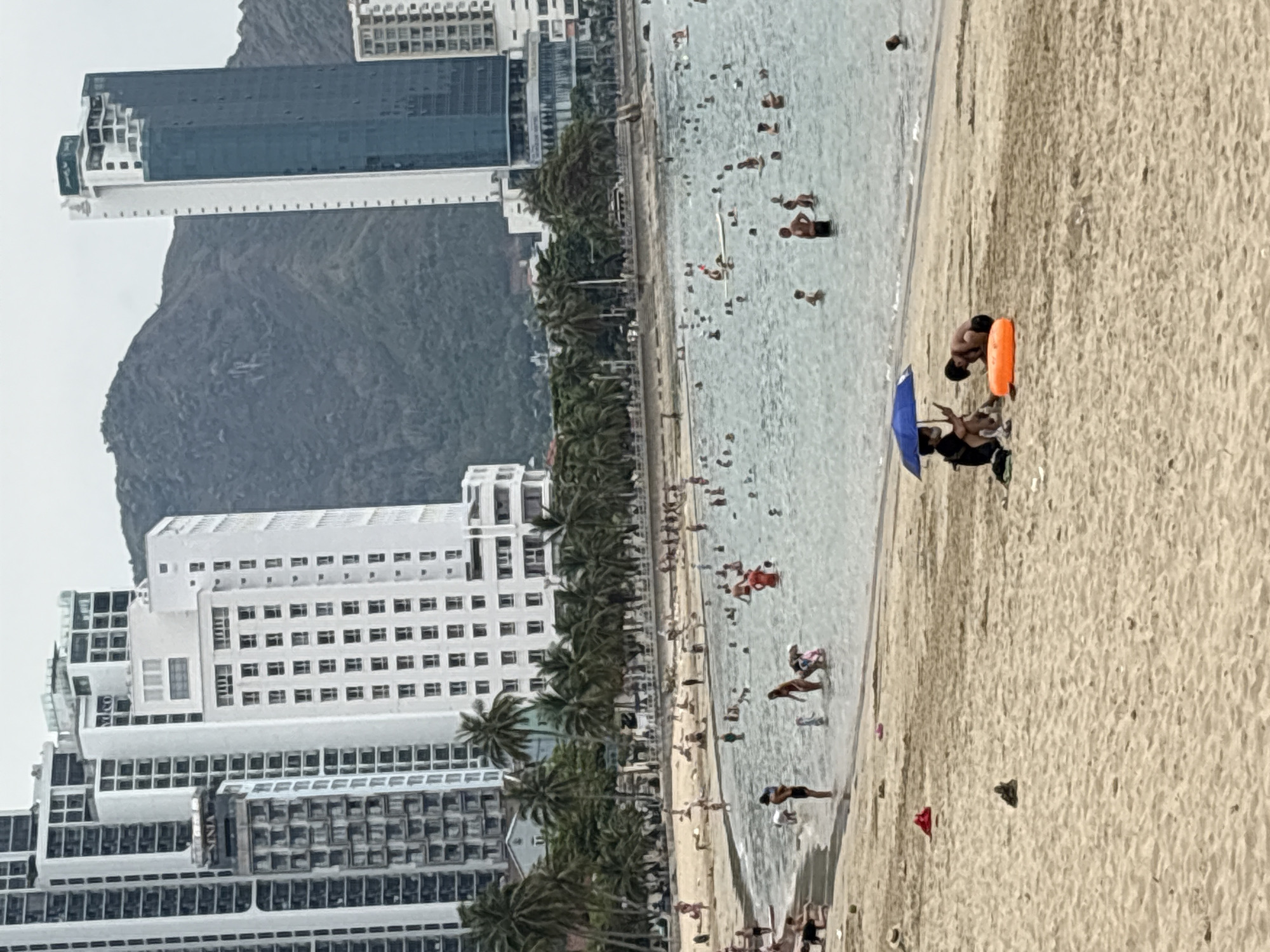 The image shows a beach in Bac Nha Trang Ward, Vietnam, with a mix of high-rise buildings and a mountain range in the background. The beach is crowded with people swimming, sunbathing, and relaxing. There are palm trees lining the beach along the shoreline. Several buildings can be seen in the background. The water appears clear and calm. There is an umbrella that is blue on the sand to the right side of the photo.