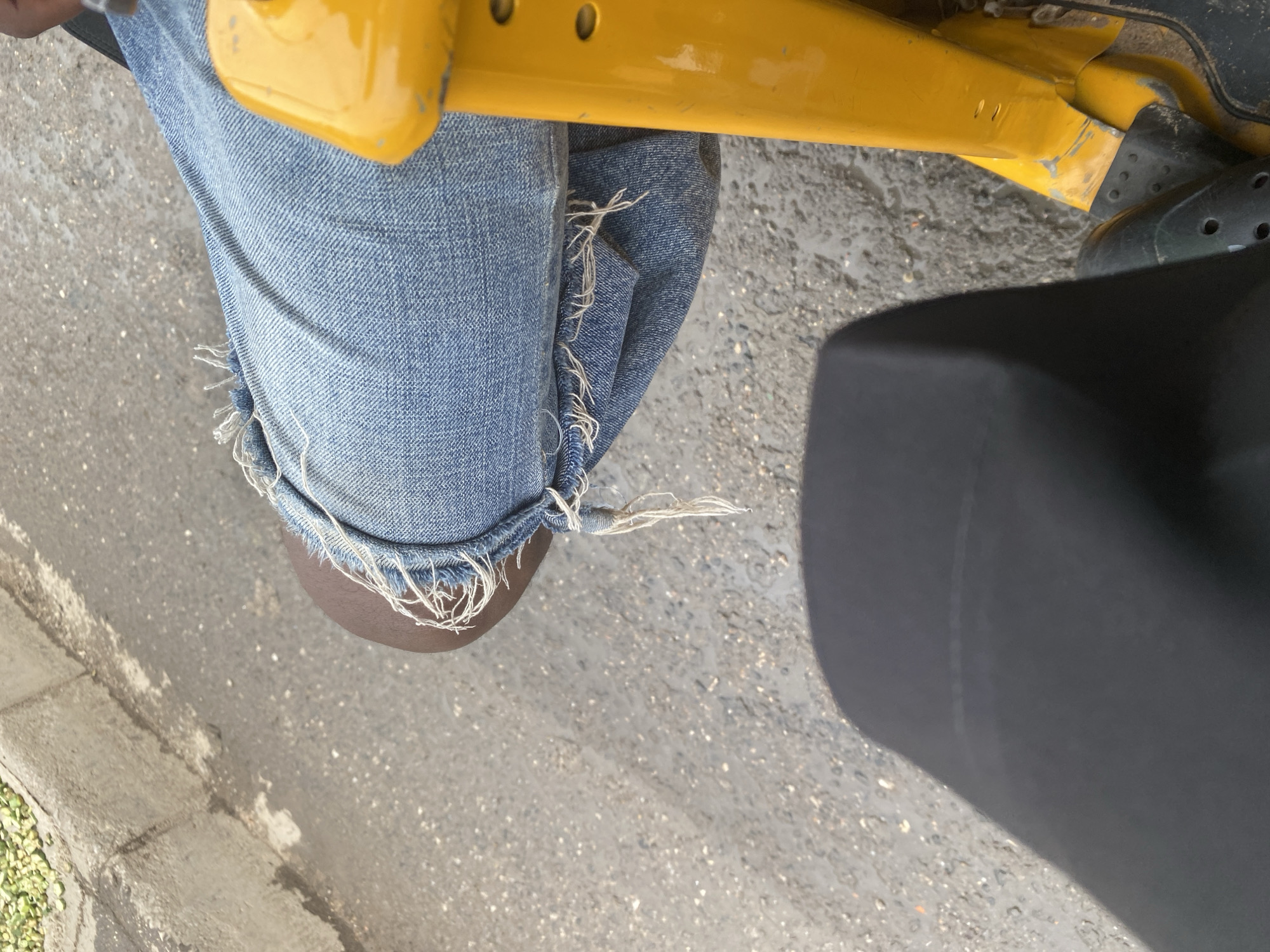 The image shows a close-up view of a person's legs and knees, wearing distressed denim jeans. The jeans have frayed edges and loose threads, particularly around the knees, suggesting a casual or worn style. The person is seated, as indicated by the angle of the legs and the presence of a dark, possibly padded, object in the foreground that could be part of a seat or vehicle. A bright yellow bar, likely a component of a vehicle or equipment, is visible across the top of the image, partially obscuring the person's upper legs and torso.
The background is a textured, greyish surface that appears to be asphalt or concrete, with some scattered pebbles and moisture, hinting at an outdoor setting, possibly a street or pathway. To the left, a rough, grey curb with some green debris is visible. The lighting suggests it is daytime, with no strong shadows indicating a specific time. There is no visible text in the image. The overall impression is a snapshot of a person in a relaxed, everyday situation, possibly waiting or traveling, in an urban or semi-urban environment in Jalingo, Nigeria.
