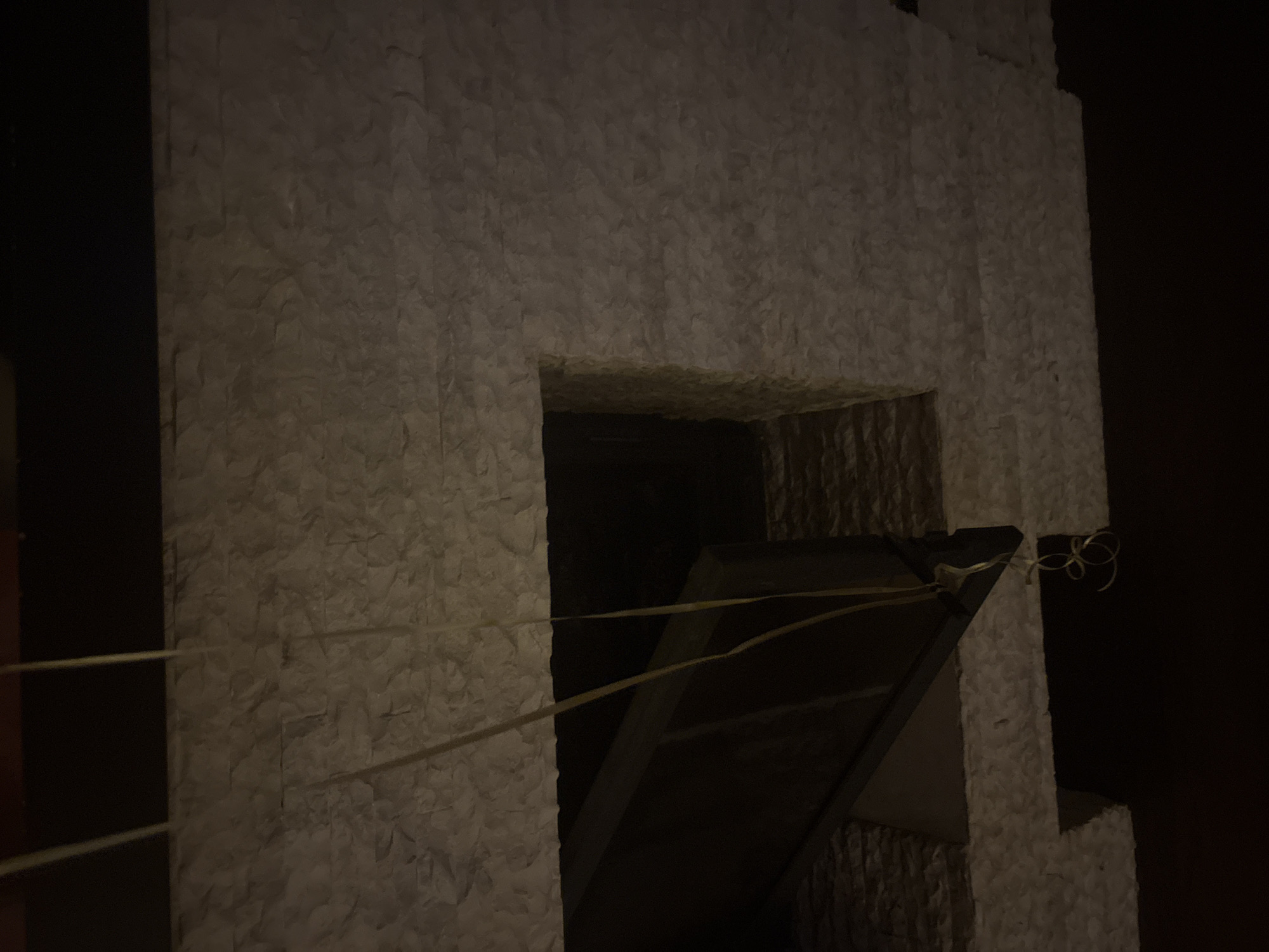 The image shows a close-up view of a textured, light-colored stone wall, possibly a facade or interior finish. A dark rectangular opening is visible in the wall, revealing a dark interior space. Two thin yellow strings or wires hang down from the top of the image, passing through the opening and disappearing into the darkness below. The stone pattern is irregular and appears to be composed of stacked rectangular pieces. The lighting is dim, casting shadows and obscuring details within the opening. There is no discernible information about a specific location in the city of Orekhovka, Russia, or any people, specific objects, or actions beyond the visual elements described.