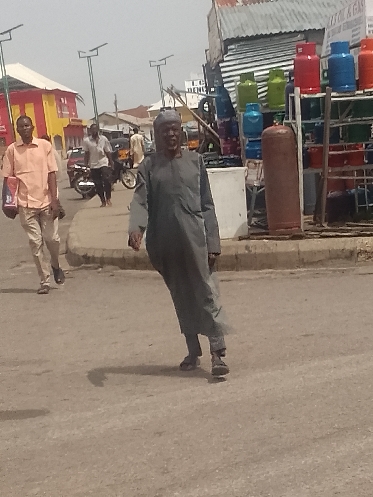 The image depicts a street scene in Pantame, Nigeria, with several individuals present. In the foreground, a man dressed in a dark gray traditional garment and a matching cap walks towards the viewer. To his left, another man in a light orange short-sleeved shirt and light-colored trousers carries a bag. Behind him, a third man in a white short-sleeved shirt and dark trousers walks away from the camera, passing a motorcycle. Further in the background, other figures and structures are visible, including buildings with colorful facades, a market stall displaying gas cylinders of various colors, and streetlights. The environment appears to be a busy urban area with paved roads and sidewalks.