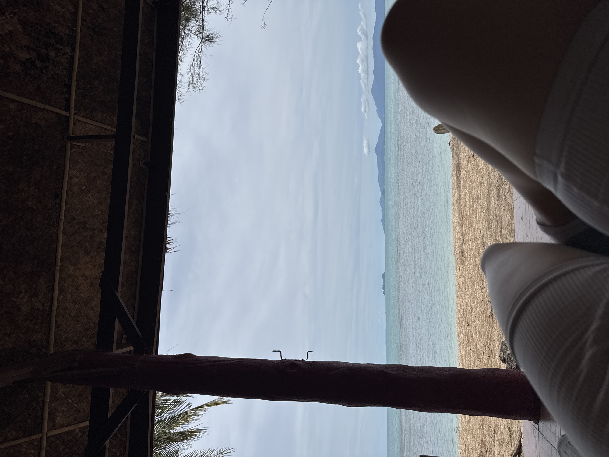 The image depicts a tranquil scene from a beachside shelter in Baan Tai, Thailand. In the foreground, a person's legs are visible, suggesting they are reclining. The shelter's thatched roof and wooden beams are in the upper portion of the frame, casting shadows. A dark, textured pillar stands to the left, partially obscuring the view of the beach and the ocean. The beach itself is sandy with scattered debris. Beyond the beach lies the calm, turquoise ocean, extending to the horizon where a range of islands and mountains are faintly visible under a cloudy sky. The overall atmosphere is one of relaxation and natural beauty.