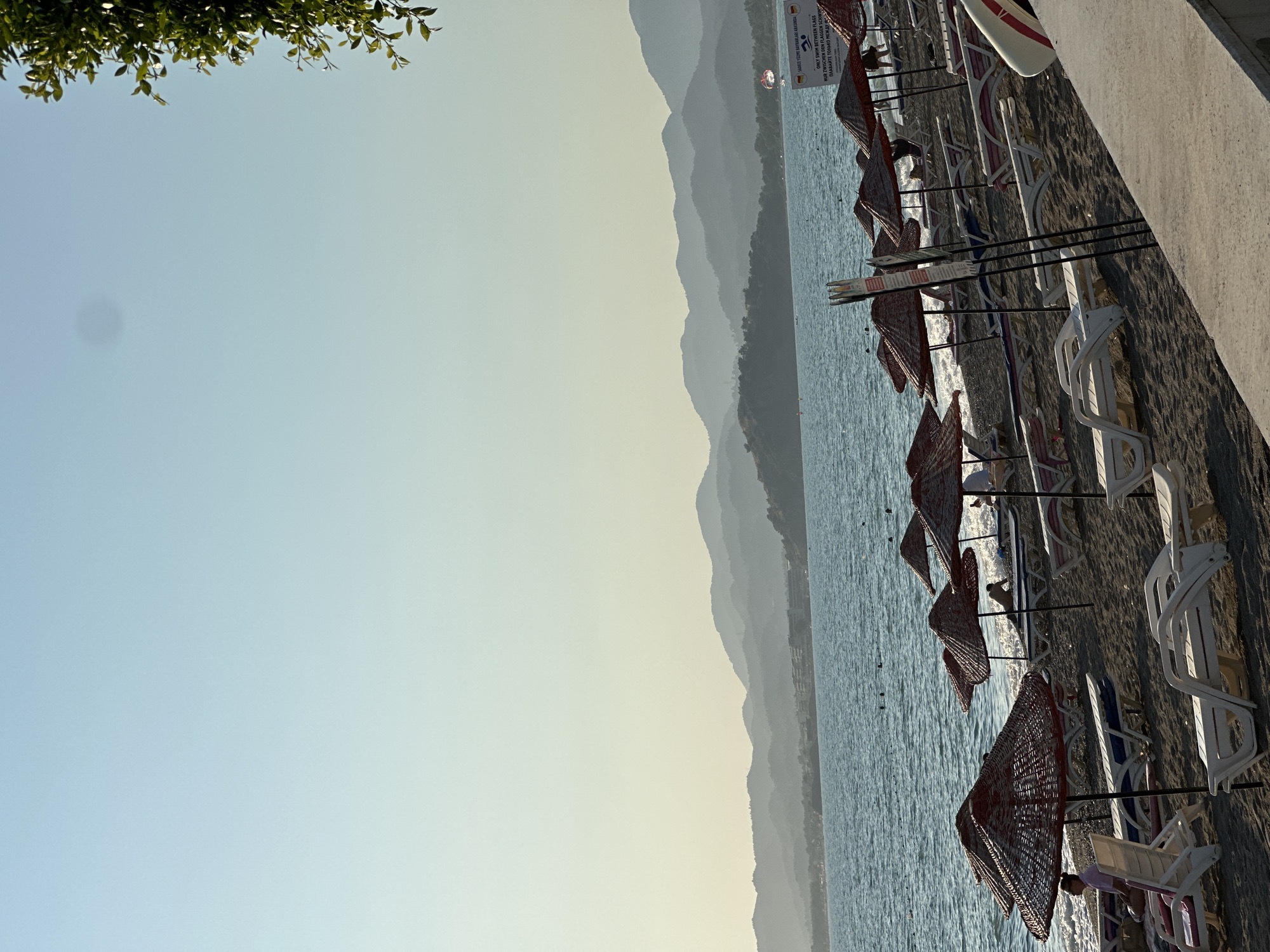 The image shows a beach scene in Fethiye, Turkey. The beach is lined with straw umbrellas and empty lounge chairs. The sea is calm and blue, with distant mountains visible in the background. The sky is clear and a pale blue. There are a few people on the beach, some of whom are sitting in lounge chairs, while others are in the water. There is a sign in the background that appears to be a set of beach rules. The overall impression is one of a peaceful and relaxing day at the beach.