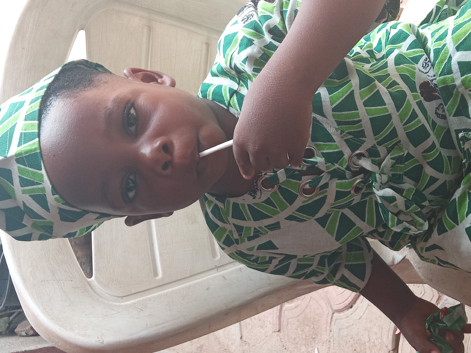 A young child, with dark skin, is the central subject of the portrait-style image. The child is wearing a vibrant green and white patterned outfit, which includes a matching head covering or cap. The outfit features decorative metal eyelets on the front, possibly for lacing. The child holds a white lollipop or stick in their mouth and looks directly at the camera with a neutral expression.
The child appears to be leaning or sitting on a light-colored, possibly white or cream, plastic chair. The chair's back is visible behind the child's head. In the lower right corner, the child's right hand is visible, holding a crumpled green object, possibly a leaf or paper. The background suggests an outdoor or semi-outdoor setting, with a reddish-brown ground surface visible in the lower portion of the image. The natural lighting indicates it is daytime. No discernible text is visible. The scene is set in Sabon Tasha, Nigeria.