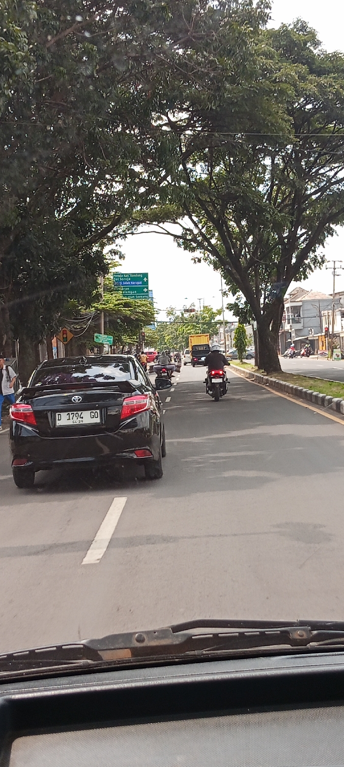 The image shows a black sedan driving down a paved road, located in Soreang, Indonesia. The road is lined with trees and a few buildings are visible on the side. There are two motorbikes driving in front of the car. The car's license plate is "D 1794 GO" and it has a windshield wiper. The image was likely taken from the perspective of another car. The sky is clear and the light is bright.  The image appears to have been taken from inside a moving vehicle and the image is out of focus towards the front.  It is a sunny day.