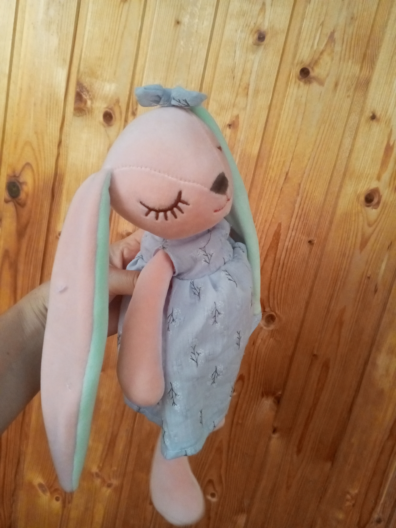 A plush toy rabbit, predominantly pink with pale green accents on its long ears, is held by a person's hand against a backdrop of a wooden wall. The rabbit is wearing a light blue dress patterned with small dark branches and leaves. It has stitched closed eyes and a small blue bow on its head. The wooden wall exhibits a natural grain and knot pattern. The image appears to be an indoor shot. The location is Chistopol, Russia.