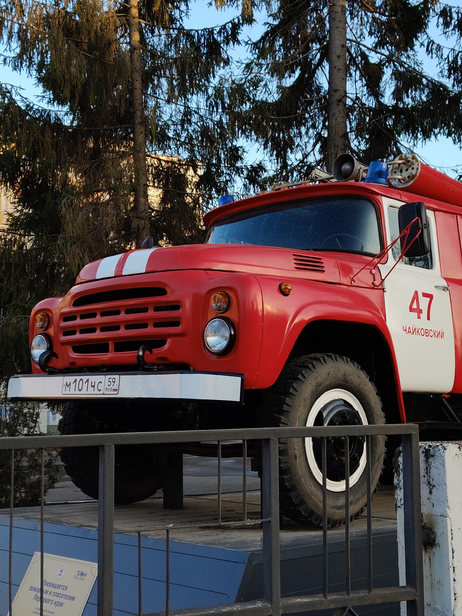 A red and white ZIL-131 fire engine with the number "47" and the word "Чайковский" (Chaikovsky) on the side is displayed outdoors. The vehicle has a white front bumper, dual white stripes on the hood, a black grill, and blue emergency lights and a siren on the roof. The license plate reads "M1014C 59 RUS". The fire engine is mounted on a concrete platform with a black metal fence. A sign is visible at the base of the platform, with text in Russian. A conifer tree is behind the vehicle, and the sky is visible in the background. The location is the city of Chaikovsky, Perm Krai, Russia.