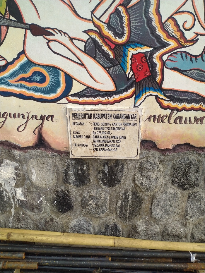 An outdoor, eye-level shot captures a vibrant mural, a project information board, and a stone wall in Karanganyar, Indonesia. The upper portion of the image is dominated by a colorful mural, likely in the style of traditional Indonesian art, featuring stylized figures with intricate patterns in shades of blue, orange, red, black, and white. Parts of the mural show human-like hands holding an object, and elaborate, wing-like or costume elements. Text from the mural, partially visible on the left, reads "gunjaya," and on the right, "melawa."
Below the mural, centered in the frame, is a rectangular white sign with black text, serving as a project information board. It details a government initiative from the "PEMERINTAH KABUPATEN KARANGANYAR" (Karanganyar Regency Government). The "KEGIATAN" (Activity/Project) includes "PEMB. GEDUNG KANTOR PERMANEN" (Permanent Office Building Construction) and "REHABILITASI STADION 45" (Rehabilitation of Stadium 45). The "BIAYA" (Cost) is listed as "Rp. 775.095.000,-" (775,095,000 Indonesian Rupiah), funded by "DANA ALOKASI UMUM (DAU)" (General Allocation Fund) for the "TAHUN ANGGARAN 2022" (Fiscal Year 2022). The "PELAKSANA" (Contractor) is "CV. CATUR MANUNGGAL KAB. KARANGANYAR."
Below the sign, a robust stone wall with irregular, dark grey rocks cemented together forms the lower half of the image. At the very bottom, several long bamboo poles and possibly some dark, cylindrical metal pipes are stacked horizontally, suggesting ongoing construction or materials storage. The lighting indicates it is daytime with clear weather. The overall scene reflects a public infrastructure project in progress or recently completed, adorned with local artistic elements.
