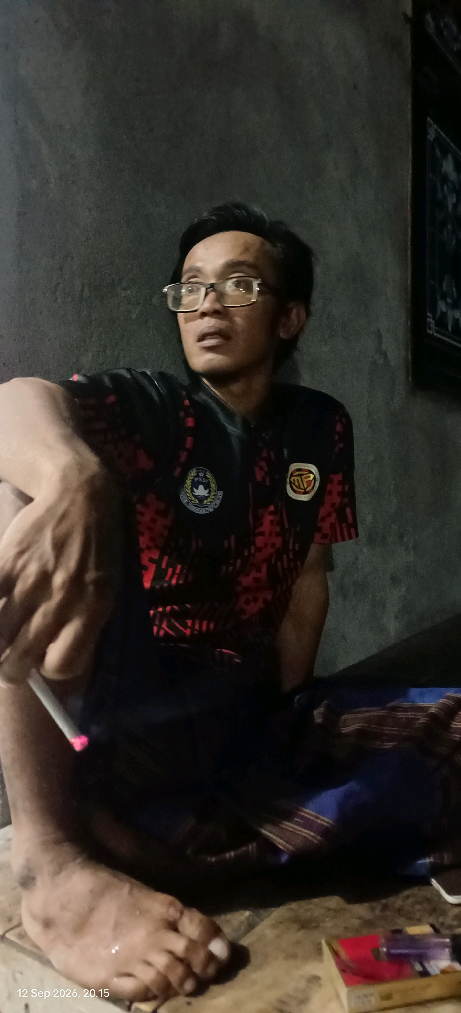 In Mojopetung, Indonesia, a man wearing glasses and a black t-shirt with a red geometric pattern sits cross-legged in a dimly lit room. The wall behind him is rough and grey. His t-shirt features two circular emblems: one on the left with text and a stylized bird, and another on the right with the letters "MP" inside a circle. He holds a lit cigarette in his left hand, the tip glowing pink, and appears to be looking upwards and to his right, with his mouth slightly open. His right leg is bent, and his left leg is partially obscured by dark fabric, possibly a sarong. His bare left foot is visible, resting on a wooden surface. A red and white object, possibly a cigarette pack, is visible in the lower right corner of the image. The timestamp "12 Sep 2026, 20.15" is overlaid on the bottom left of the image.