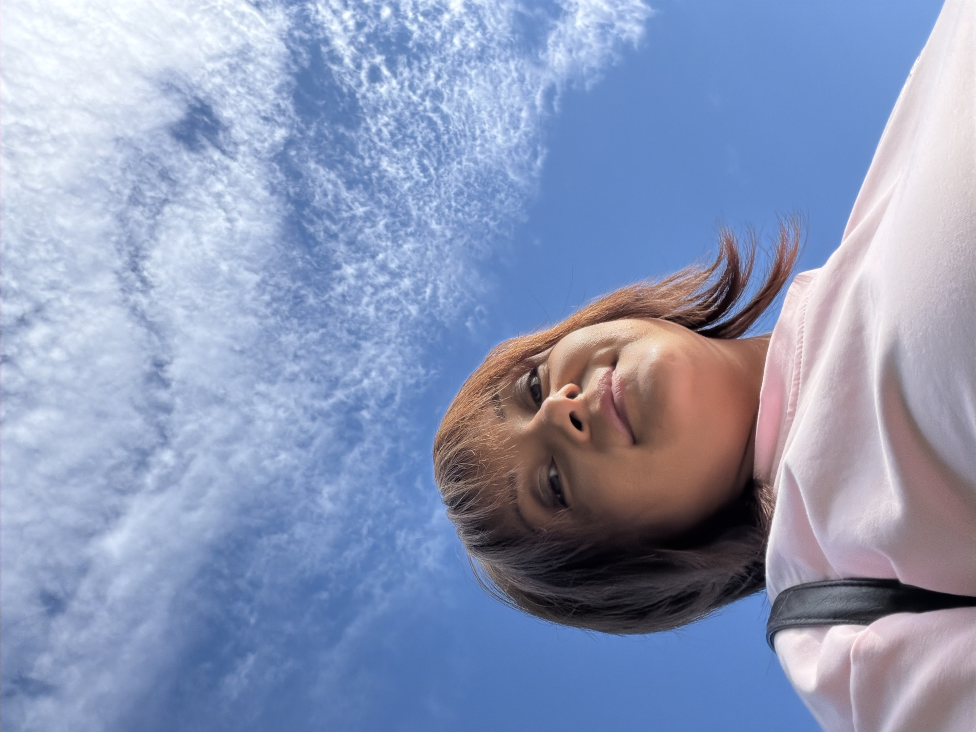 A low-angle, close-up selfie shows a person's face from the chin up, with the sky and clouds filling the rest of the frame. The person has brown hair styled with bangs and is looking up and towards the camera. They are wearing a light pink top with a black strap visible on the right side. The sky is a clear blue with wispy white clouds scattered across it, suggesting it is daytime with fair weather. The perspective is from below, as if the person is lying down or holding the camera above their head. The overall mood appears relaxed and possibly playful. There is no visible text in the image.