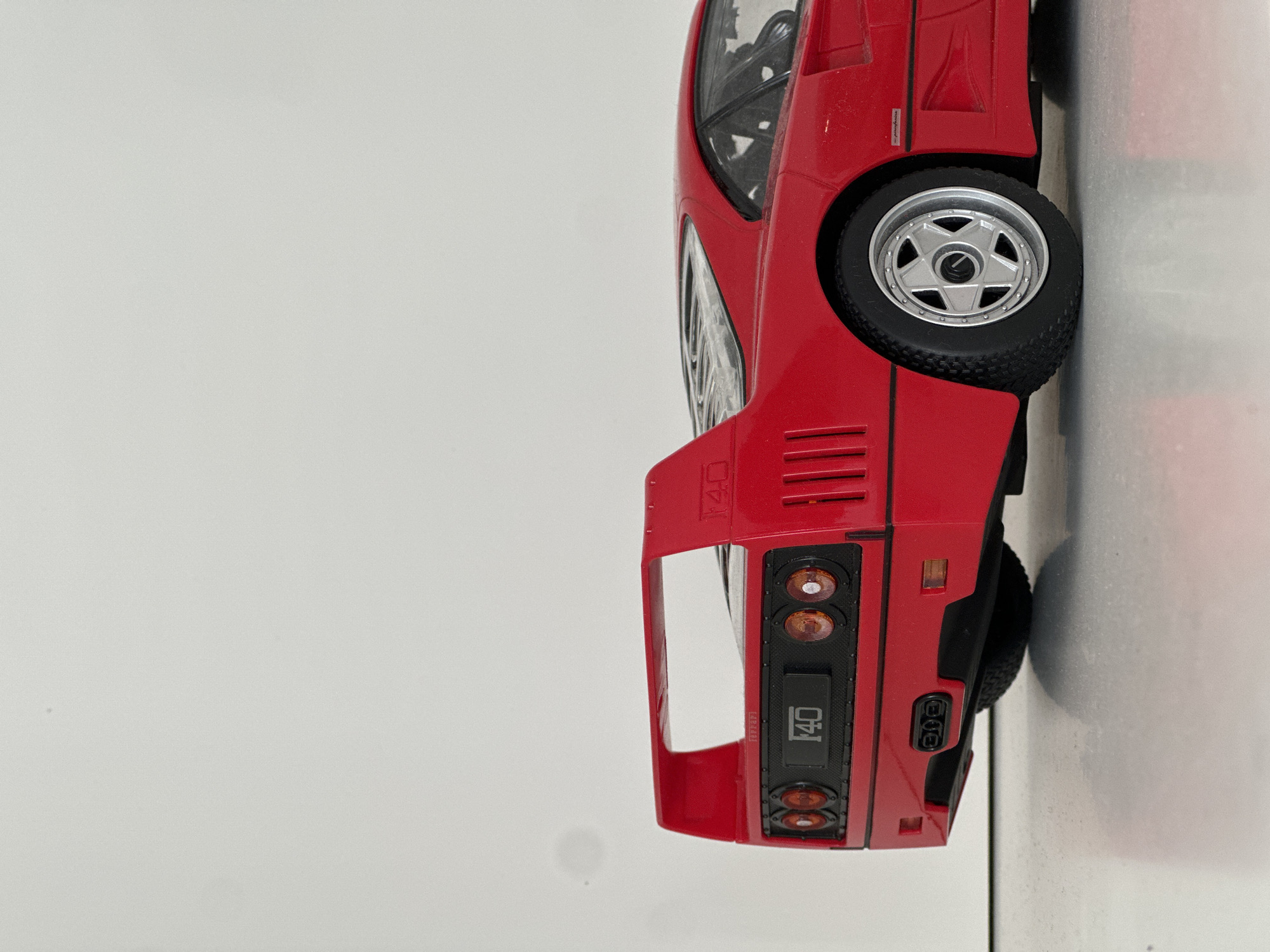 A red model car, possibly a Ferrari F40, is positioned on a white surface. The car is oriented vertically in the frame, showing the rear and side profile. The rear features visible taillights, the "F40" logo, and the spoiler. The side shows a portion of the wheel, a side vent, and the window. The background is a plain white wall. There is no indication of Milan, Italy or any people present.