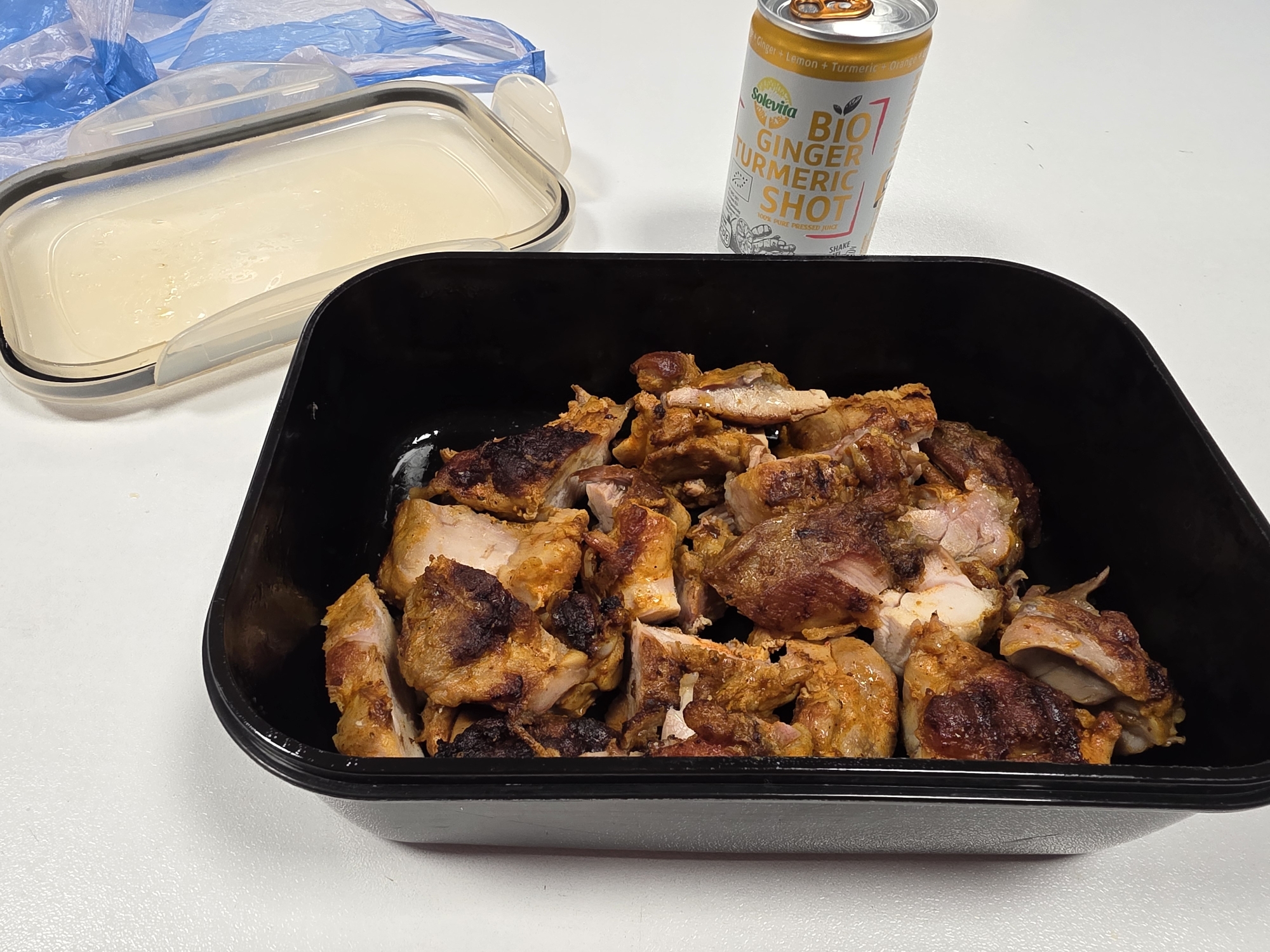 The image presents a lunch setting on a white surface, featuring a partially opened container of what appears to be a creamy sauce to the left. A can of "Solevita BIO GINGER TURMERIC SHOT" is positioned to the right, and a black container filled with diced, cooked meat occupies the foreground. A blue plastic bag is visible in the background on the left side. The scene appears to be indoors. There is no information in the photo to indicate this is Plovdiv, Bulgaria.