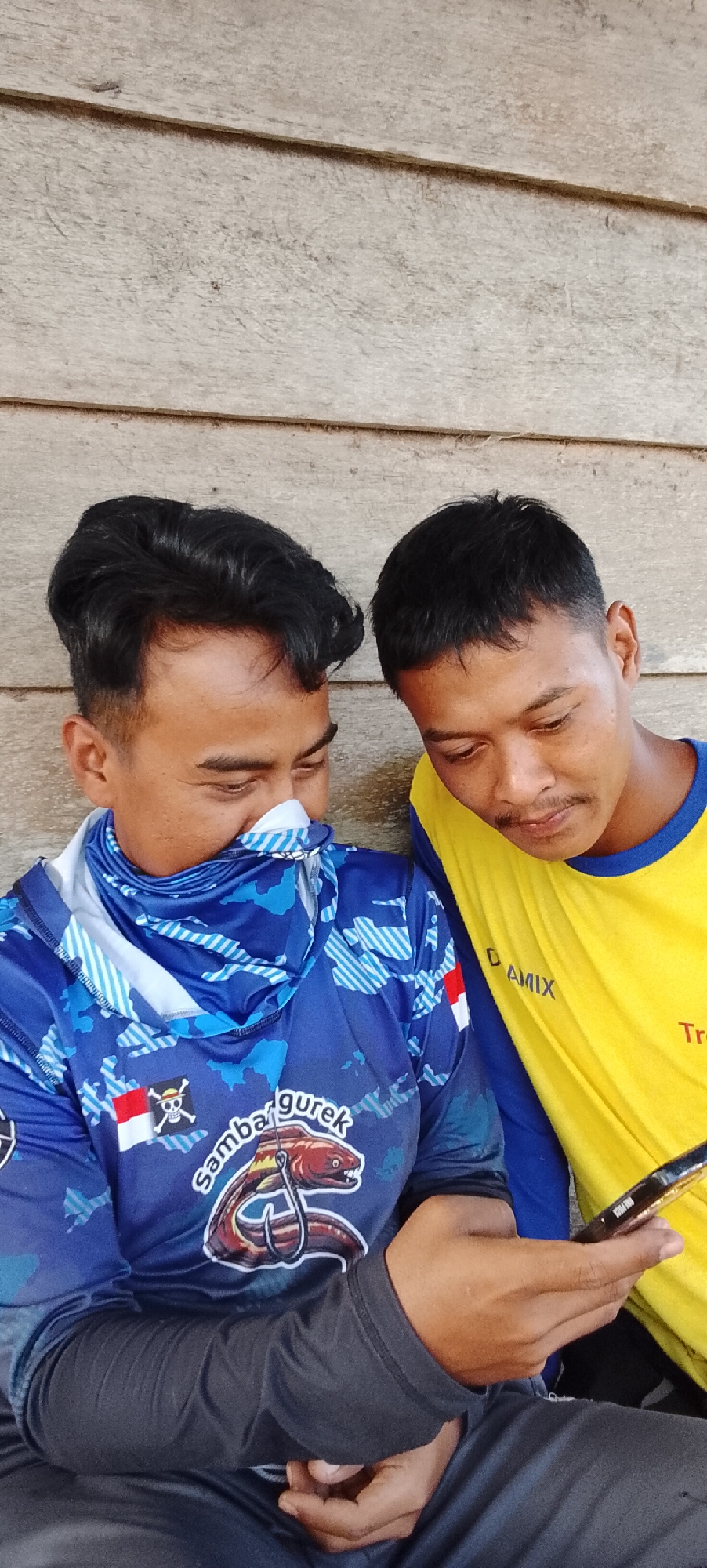 Two men are sitting close together, leaning against a weathered wooden wall. The man on the left wears a blue long-sleeved shirt with a white and blue patterned neck gaiter covering the lower half of his face. The shirt features a prominent graphic of an eel on a fishing hook, along with Indonesian flags and what appears to be text. The man on the right is wearing a yellow long-sleeved shirt with blue trim. Both men are looking down at a dark-colored mobile phone held by the man on the right, suggesting they are engaged in a shared activity or conversation on the device. The background is a simple, horizontally planked wooden wall, typical of outdoor structures in rural or semi-rural settings. The overall lighting suggests it is daytime. The image appears to be taken outdoors, likely in a village or a less urbanized area. Based on the context of the graphic on the shirt and the general aesthetic, the scene is plausibly located in a coastal or fishing community in Padang Dalom, Indonesia.