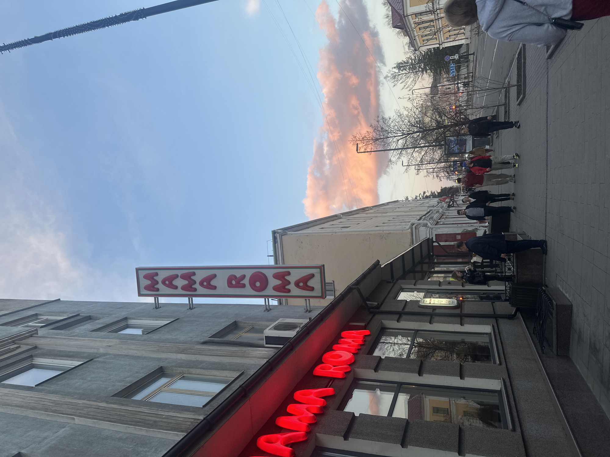 The image depicts a street scene in Krasnoyarsk, Russia, at dusk. In the foreground is a building with a large, illuminated red sign reading "MAMA ROMA." Above the building is another sign with the same name, but in a different style. The sky transitions from blue to orange, with a prominent cloud formation. Several people are walking along the sidewalk. Other buildings and trees are visible in the background. A few windows on the building reflect sunlight. A pole runs horizontally across the top of the image.