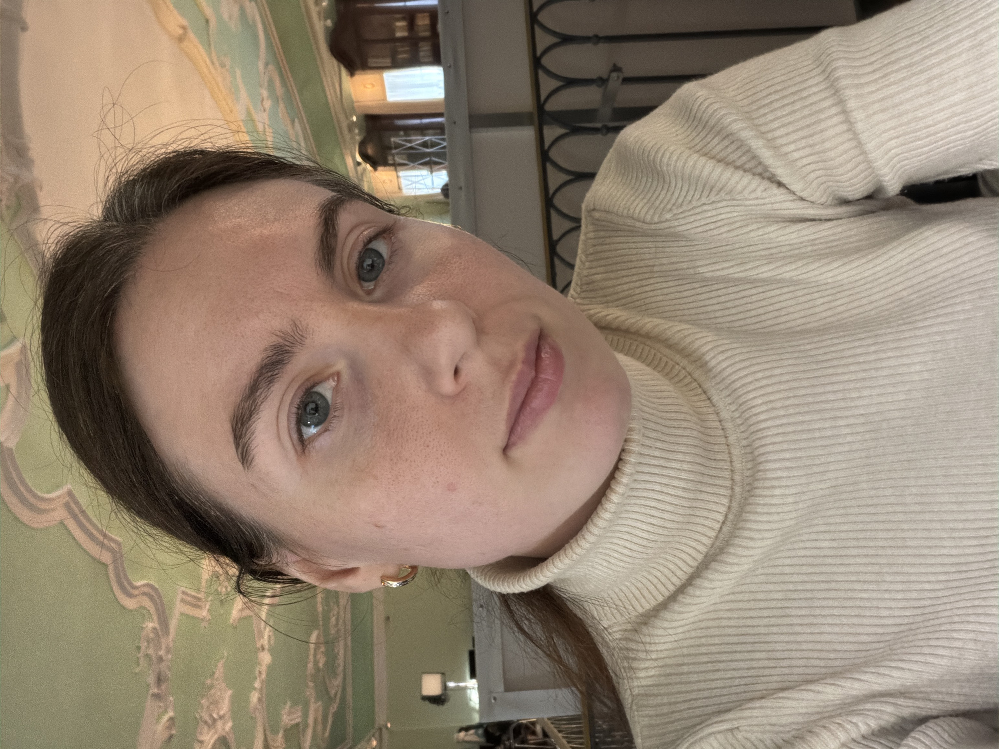 A close-up, slightly angled shot of a fair-skinned woman with dark hair and blue-gray eyes. She is wearing a light beige turtleneck sweater. The background appears to be an ornate, light green and off-white wall with classical-style decorations, suggesting an interior, potentially in Saint Petersburg, Russia, given the architectural style. A dark, decorative railing is vaguely visible behind her, further suggesting an indoor location, possibly a museum or historical building. The image appears to be taken indoors under natural light.