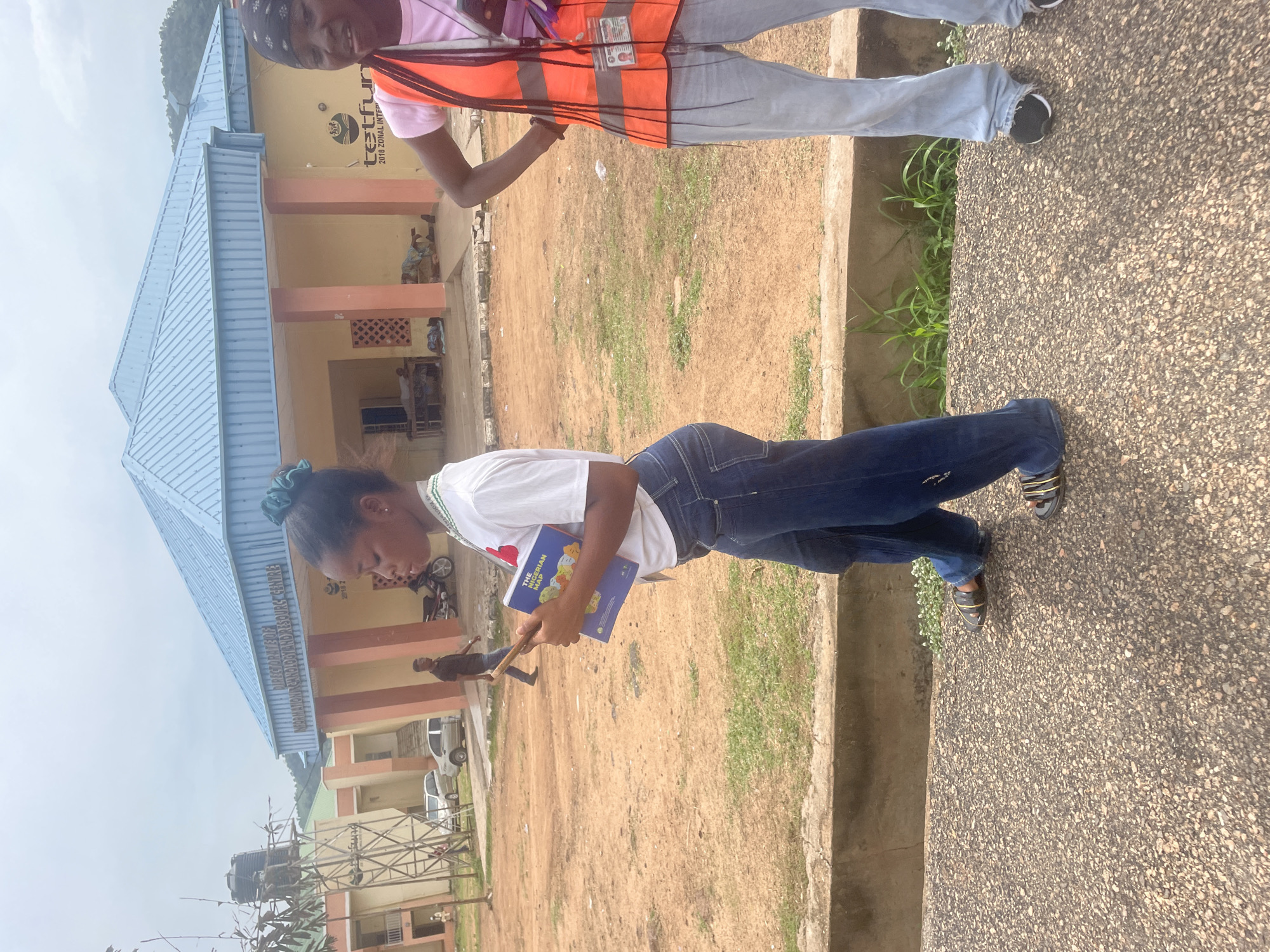 In Lokoja, Nigeria, two young women are standing on an asphalt path in front of a light yellow building with a blue metal roof. The building has pillars painted orange. A sign on the building reads "DIRECTORATE OF INFORMATION TECHNOLOGY AND RESOURCE CENTRE." One woman is wearing a white t-shirt, blue jeans, and sandals. She is holding a blue book titled "The Nigerian Map" and a mobile phone. Her hair is styled in a teal scrunchie. The second woman is wearing a pink t-shirt, a bright orange safety vest, and light blue jeans. She is also wearing black athletic shoes. The ground is covered in reddish-brown dirt and patches of green grass. In the background, there are trees, a water tower, and other buildings. A man is walking in the background, and a motorcycle is parked near the building. The sky is overcast.
