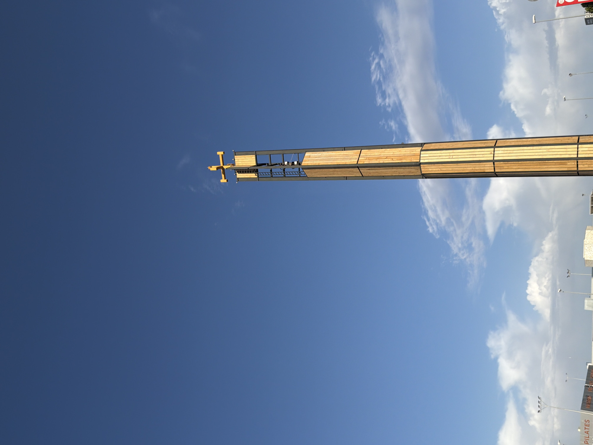 This is a low-angle, outdoor, full-shot photograph of a tall, modern tower with a cross at its apex, located in Majadahonda, Spain. The tower, which is made of wood and metal, features a ladder visible on its side. The sky is a clear, deep blue with scattered white clouds. In the foreground, a sign that reads "PILATES" is partially visible, along with streetlights and what appear to be other buildings. The scene is captured during daylight.